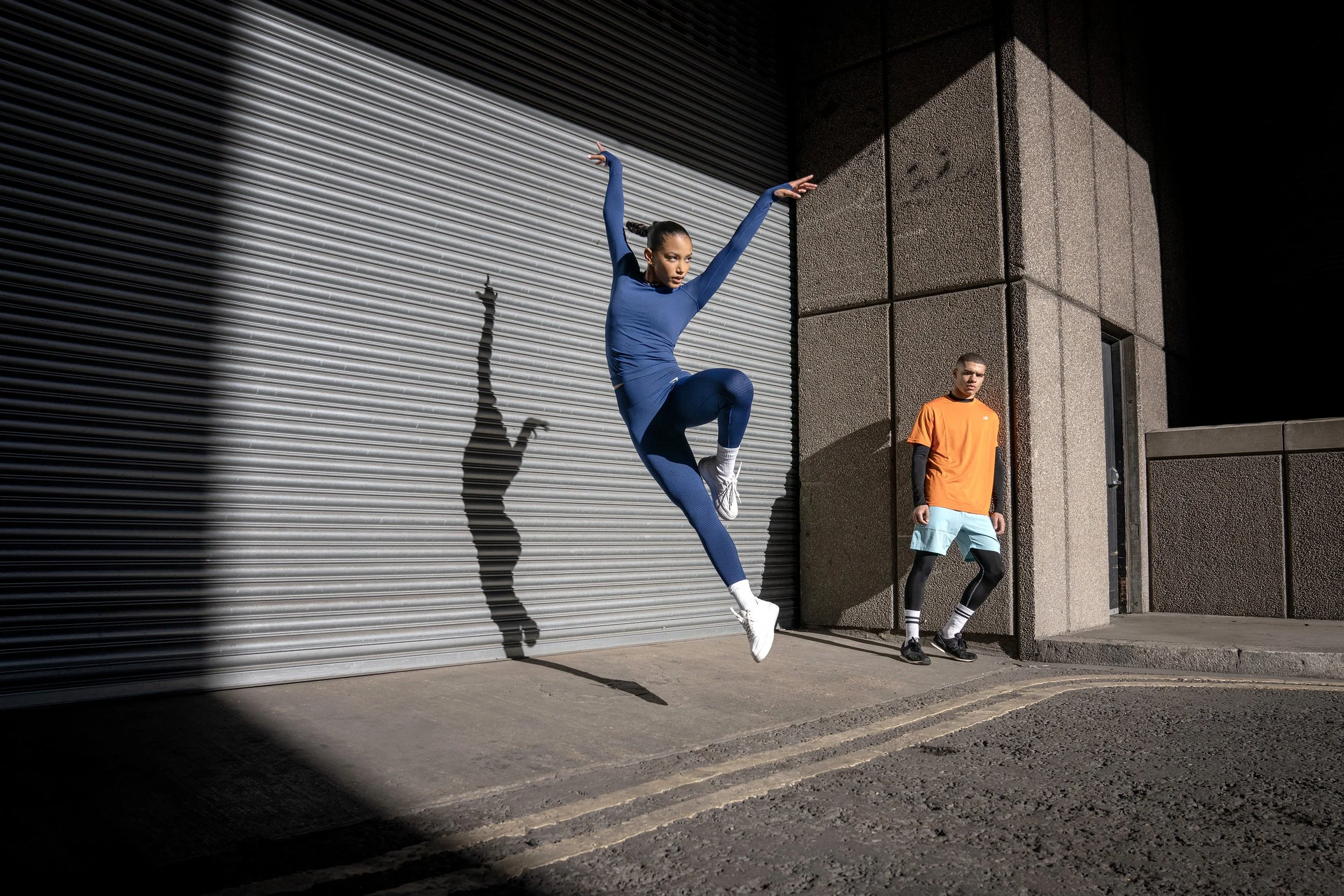 A woman in a blue A young woman leaps gracefully into the air, her shadow cast agains the wall by the harsh sunlight in a brutalist urban scene, while behind her a young man leans on a wall. dynamic sports photography by Steve Bright
