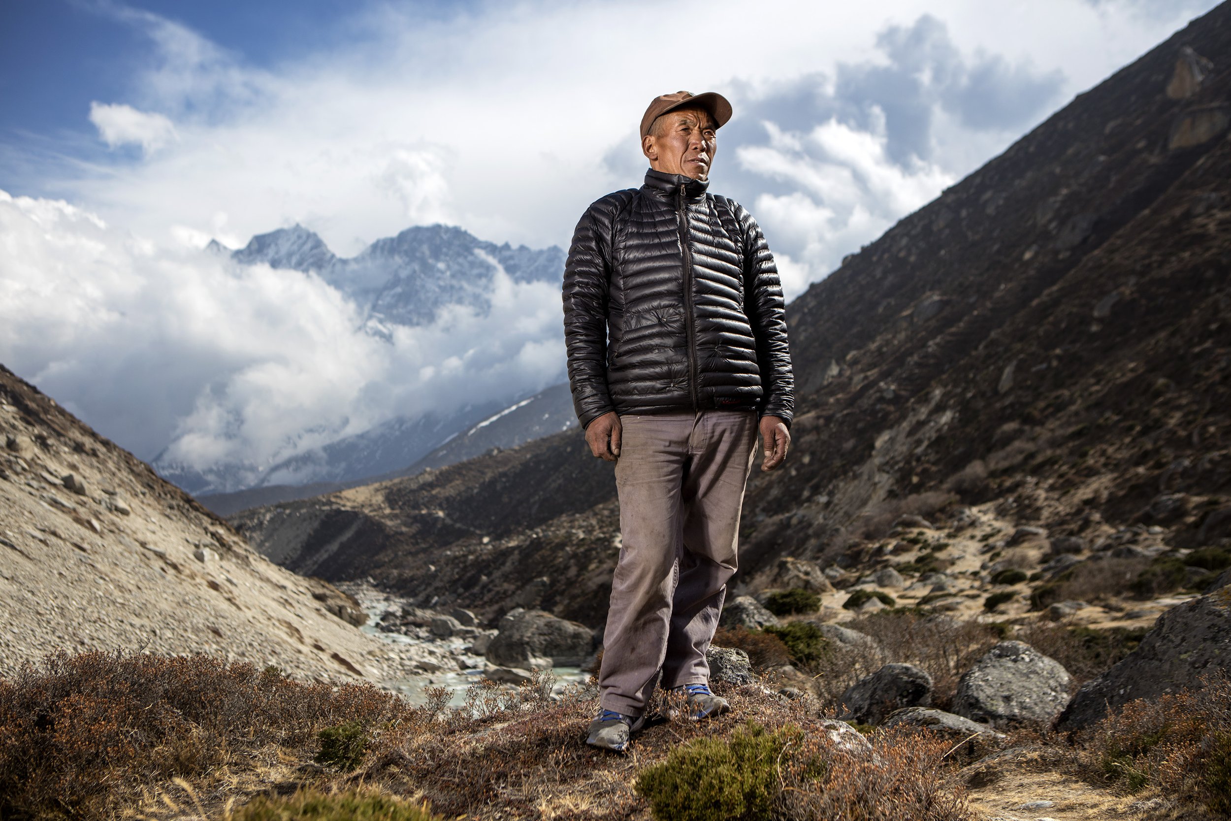 An elderly Nepalese sherpa stands in a rocky valley in the Nepalese Himalayas, wearing a baseball cap and a down jacket. commercial portrait photography by Steve Bright