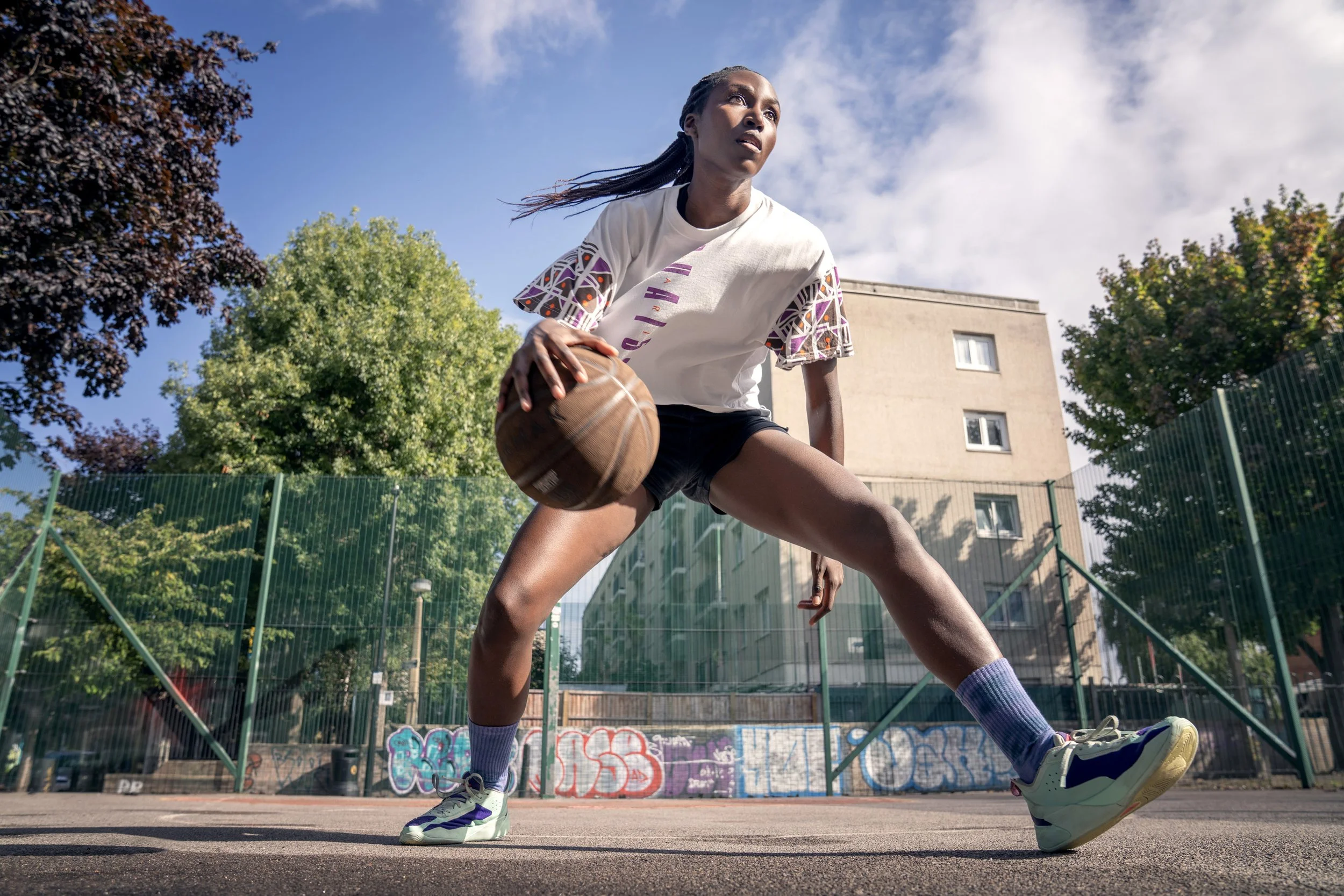 A woman playing basketball on an outdoor court, holding a basketball with a stance ready for action, with trees, graffiti, a building, and a fence in the background on a sunny day. Dynamic sports portrait photography by Steve Bright