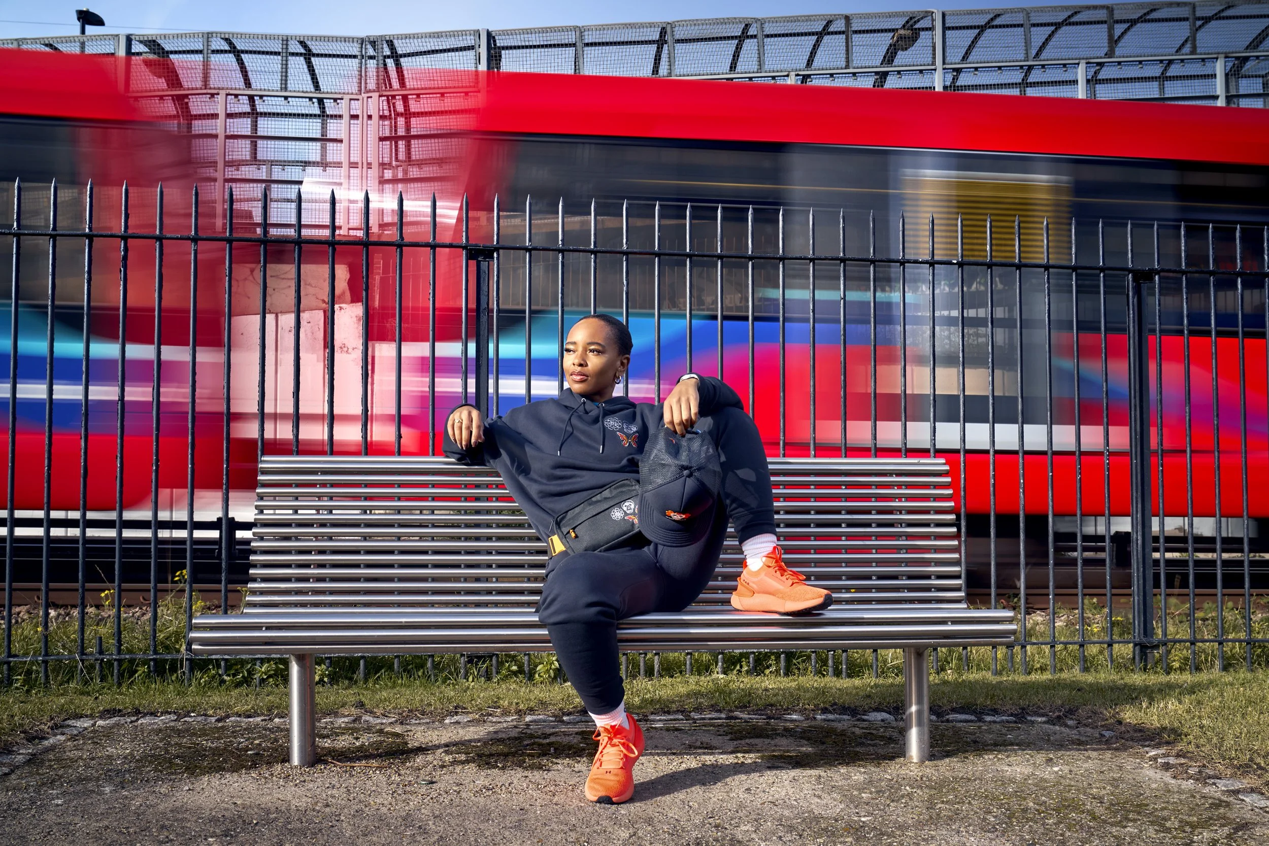 A young woman in a tracksuit and orange trainers sits on a bench in a relaxed pose, while a DLR train goes past behind, it's carriages motion blurred. commercial portrait photography by Steve Bright