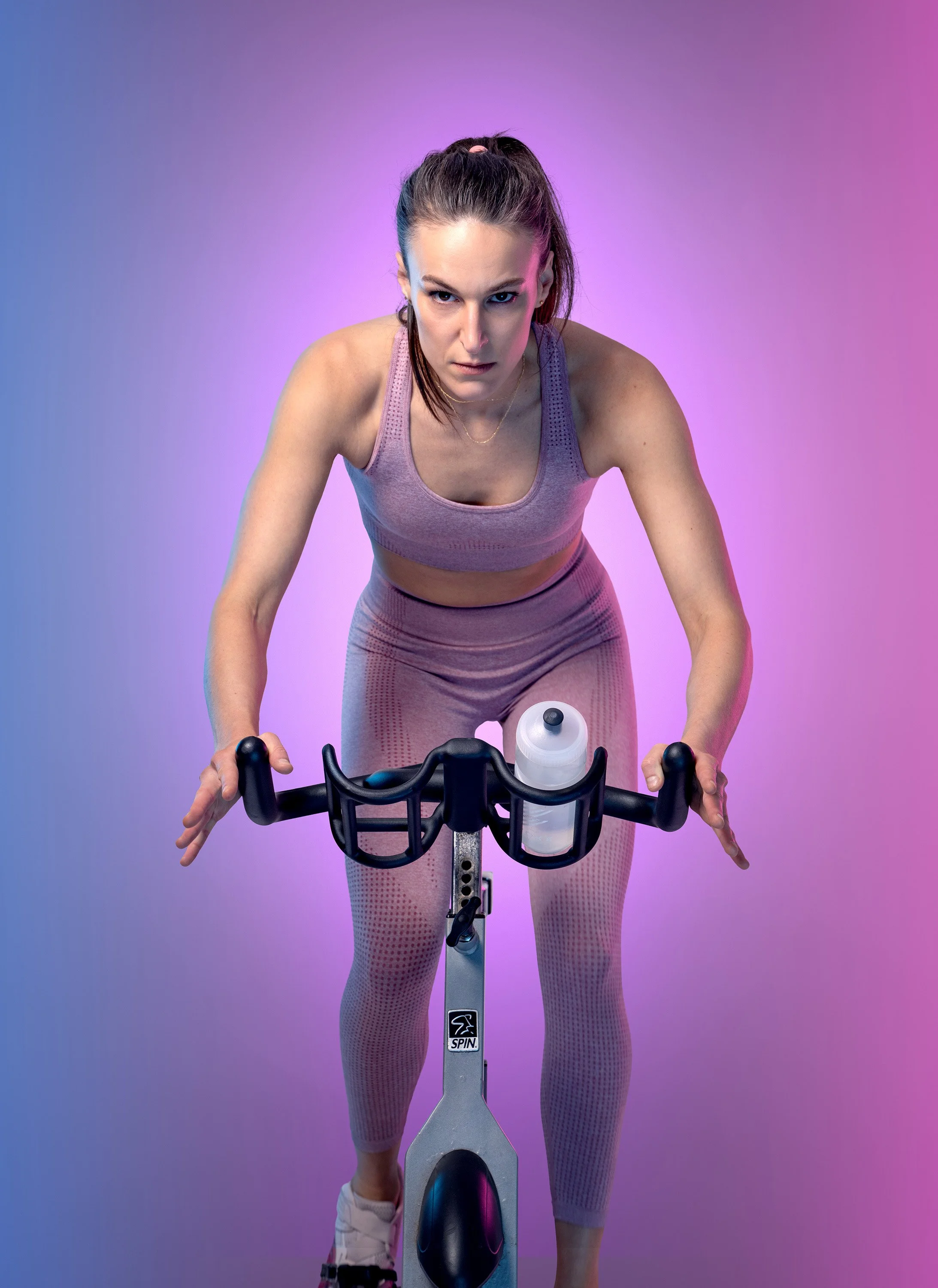 Poster image for the theatre show Spin. Actor Kate Sumpter sits on a spin bike staring at the camera, on a blue and pink background. dynamic entertainment portrait photography by Steve Bright London
