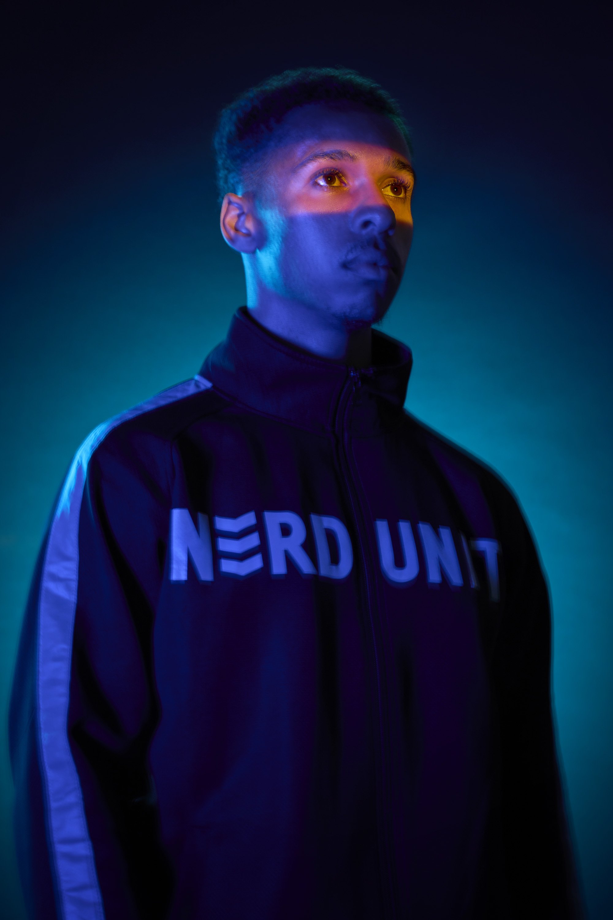 A young man in a dark shadowy image stares ahead, the only bright light a strip of orange light that lights up his eyes. He wears a Nerd Unit track top
