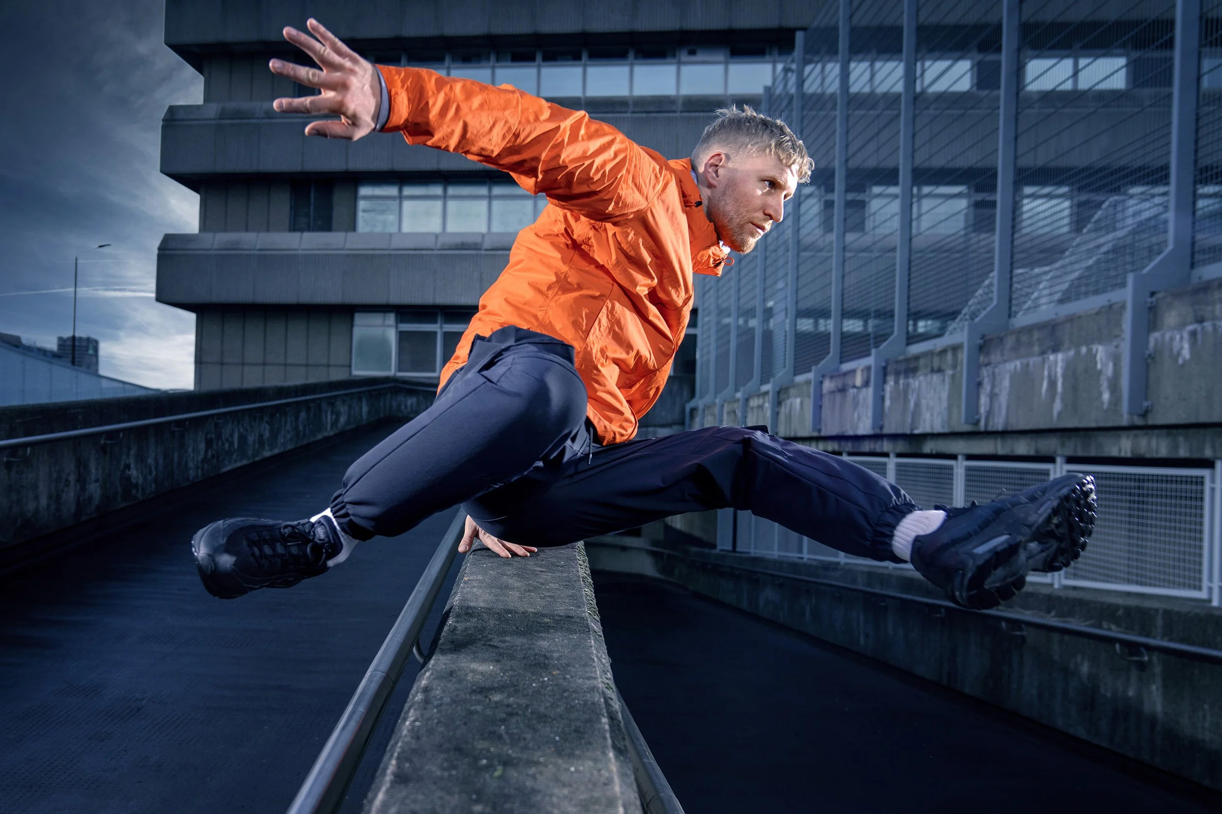Man in an orange jacket vaulting over a ledge with one hand, performing a parkour move in an urban environment. dynamic sports  portrait photography by Steve Bright
