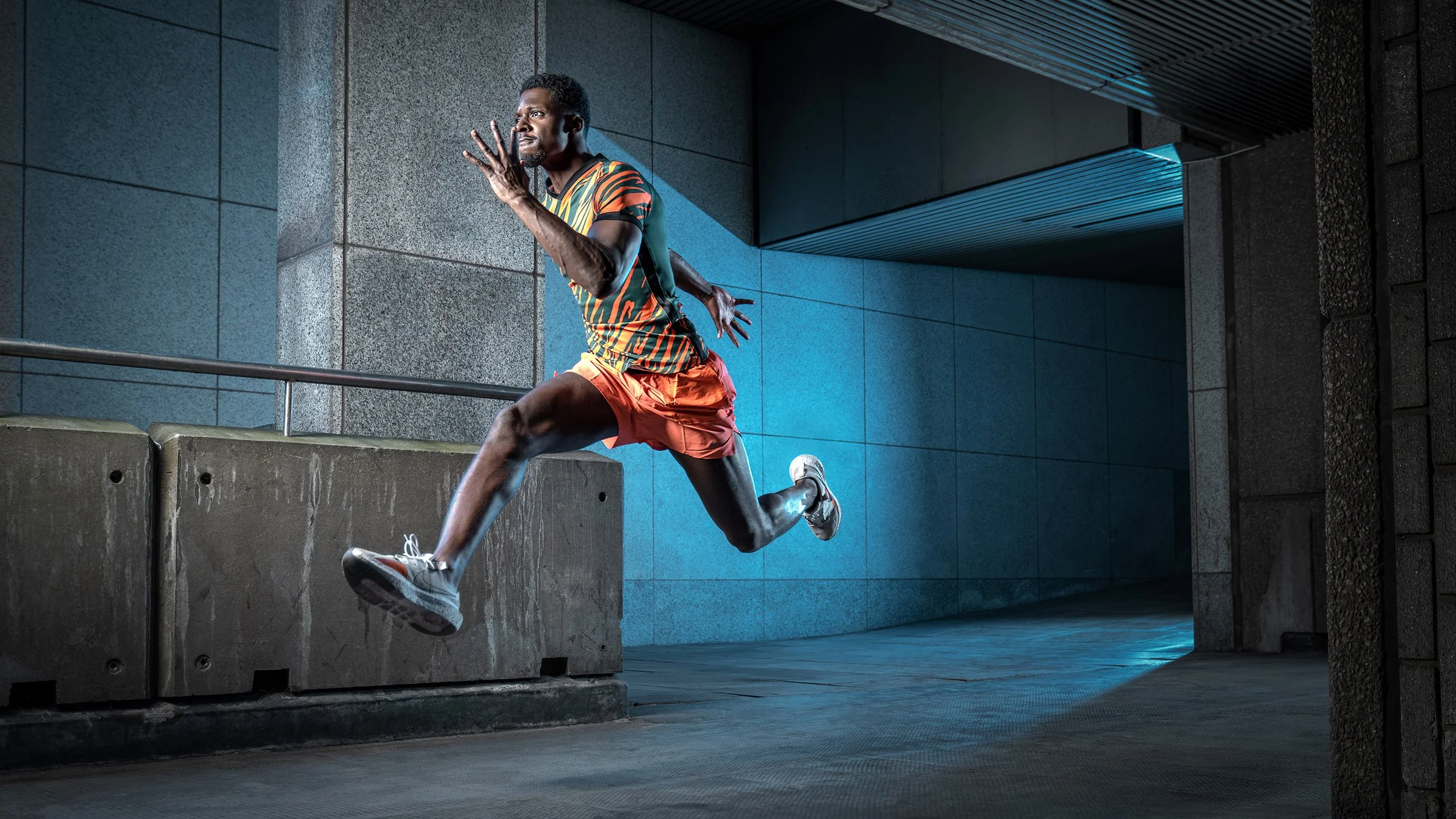A male athlete runs at full speed down an outdoor, urban walkway, lit from behind with blue light.