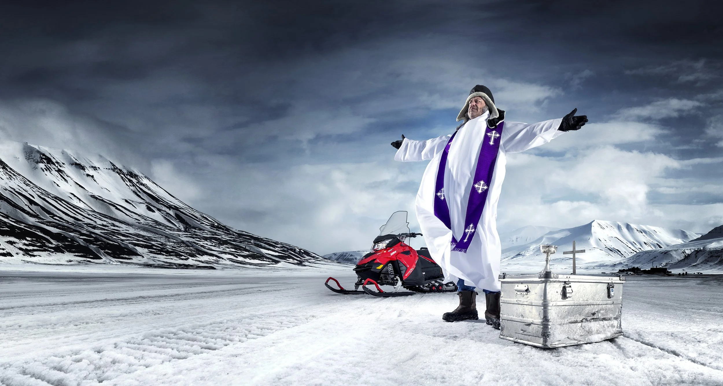 An anglican vicar stands on the snowy plains of Longyearbyen, arms outstretched, with his communion items in a metal flight case and a snowmobile behind him. commercial portrait photography by Steve Bright