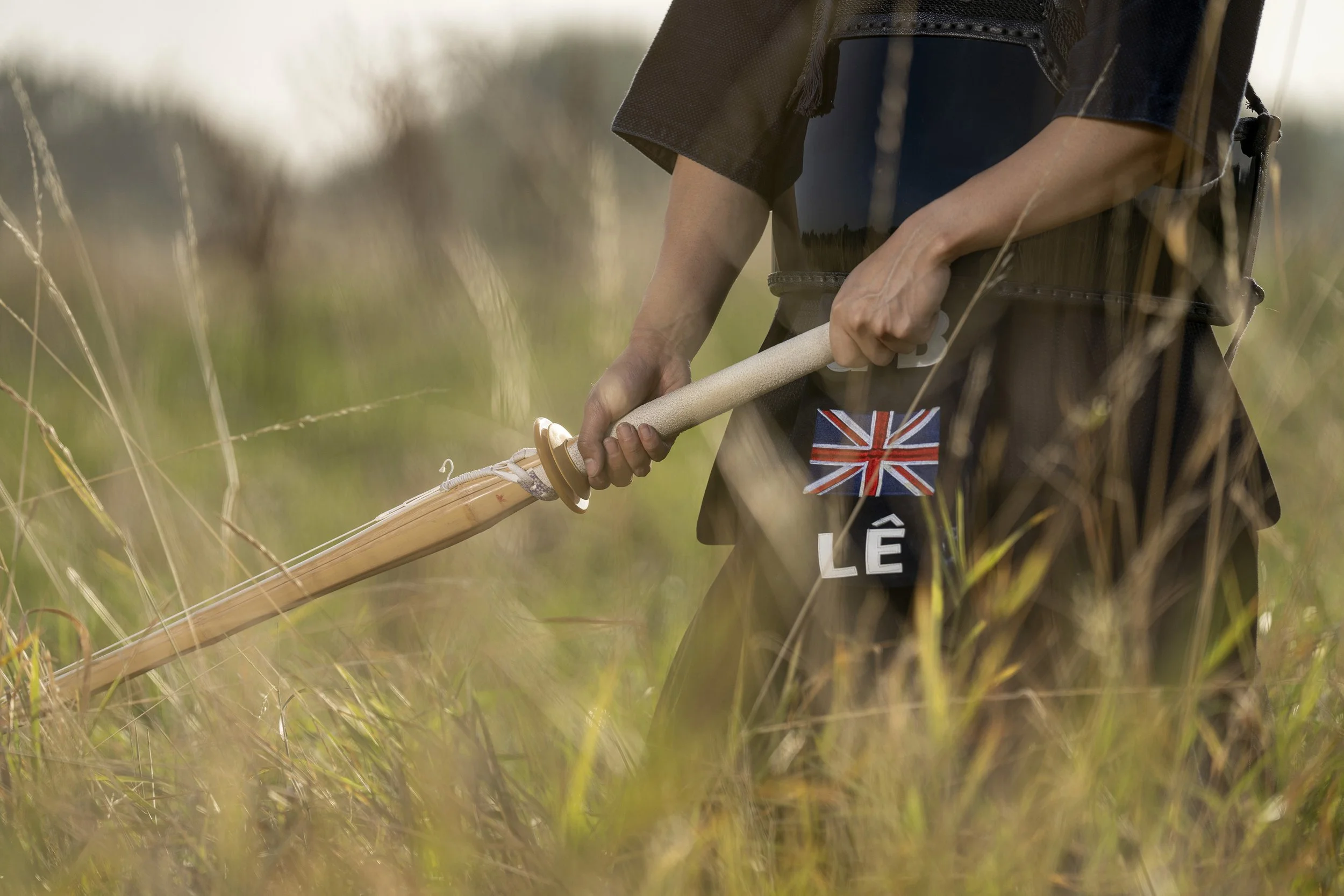 Person in traditional kendo armor, holding a wooden sword, standing in a grassy field. dynamic sports portrait photography by Steve Bright