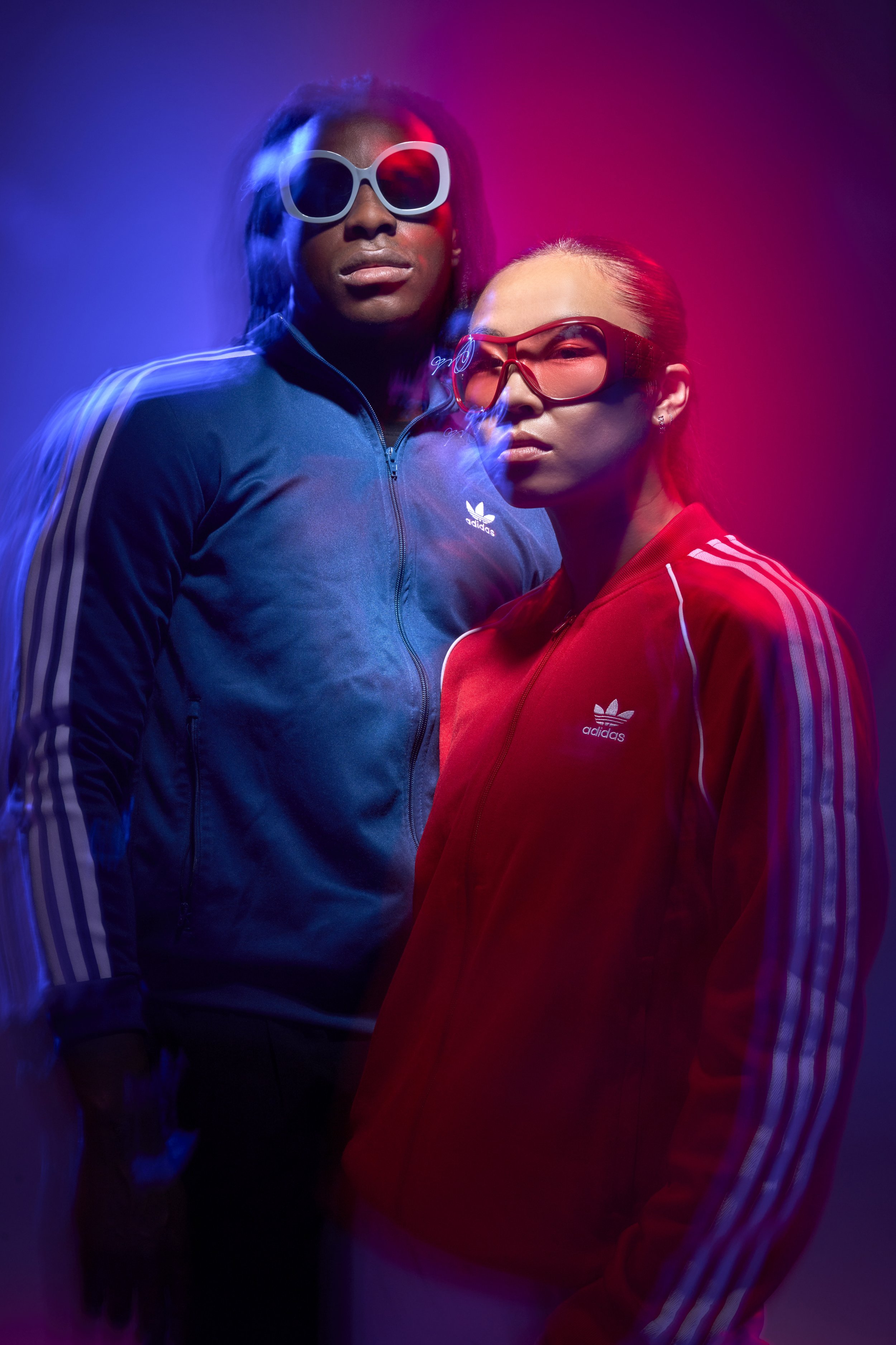 A young couple wearing red and blue adidas tracksuits and matching sunglasses look at the camera surrounded by blurry pink and blue light