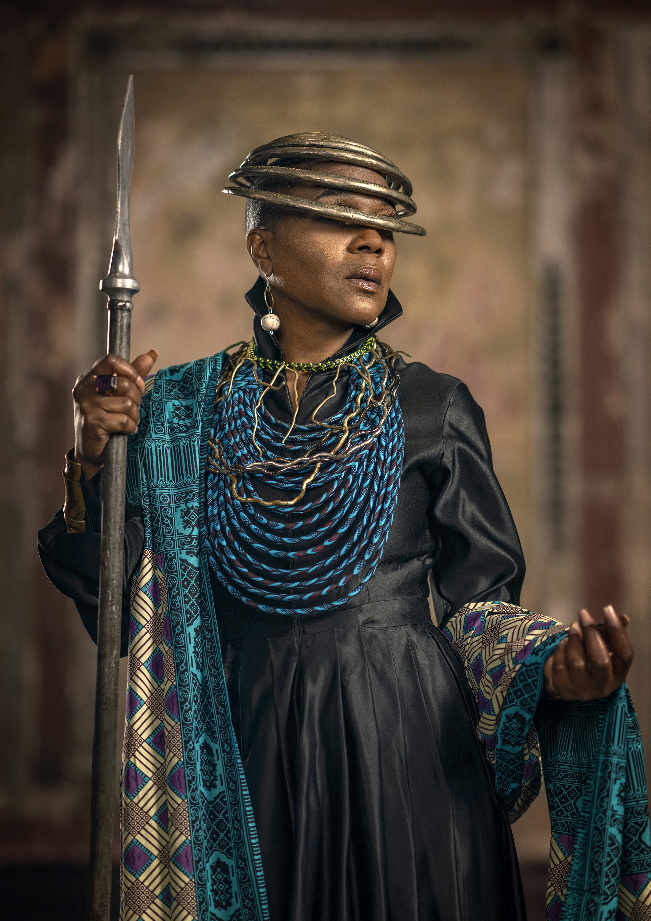 A bald Black woman with a metal headdress poses holding a spear, in a modern Afrofuturist version of a royal portrait. commercial portrait photography by Steve Bright