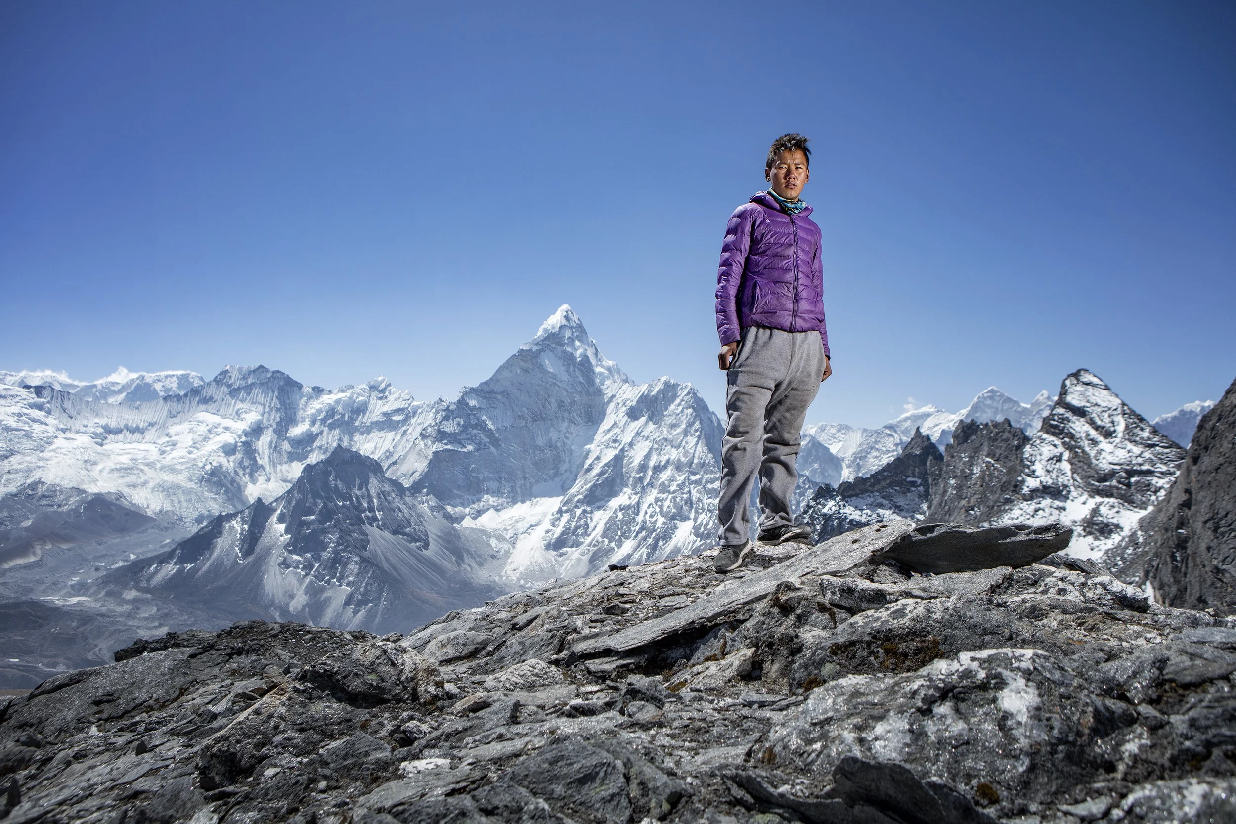 A young Nepalese sherpa stands on the peak of a mountain in the Himalayas, other snowy peaks in the background. dynamic sports portrait photography by Steve Bright