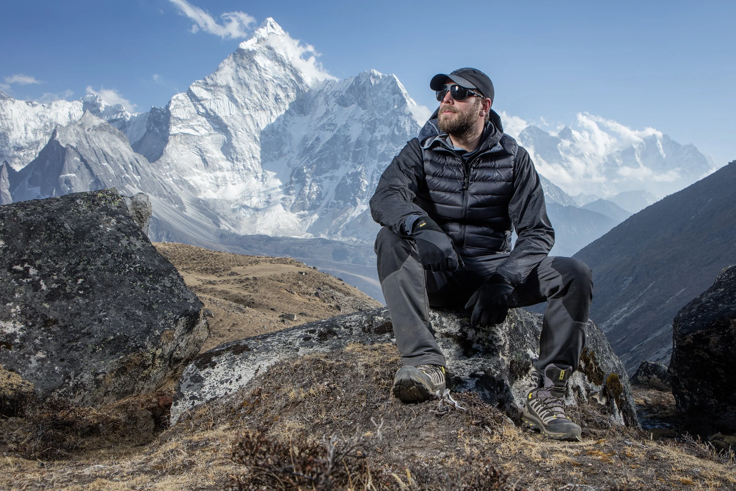 A male climber with a beard, wearing trekking clothin, sits on a rock with a spectacular range of mountains behind him. commercial portrait photography by Steve Bright