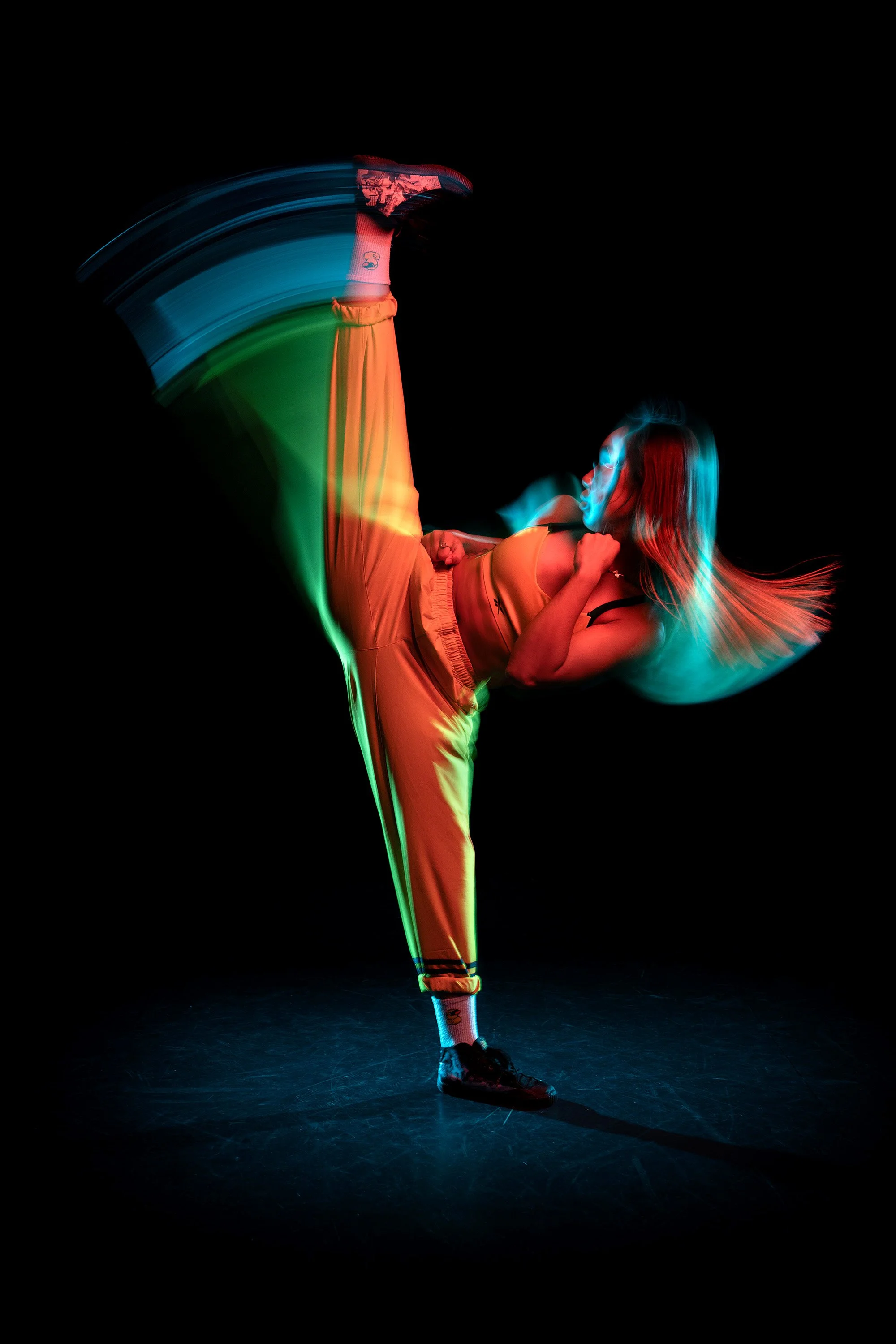 A female martial artist performing a high kick  against a black background, illuminated with colorful and blurred lights creating a vibrant, artistic portrait. dynamic sports  portrait photography by Steve Bright