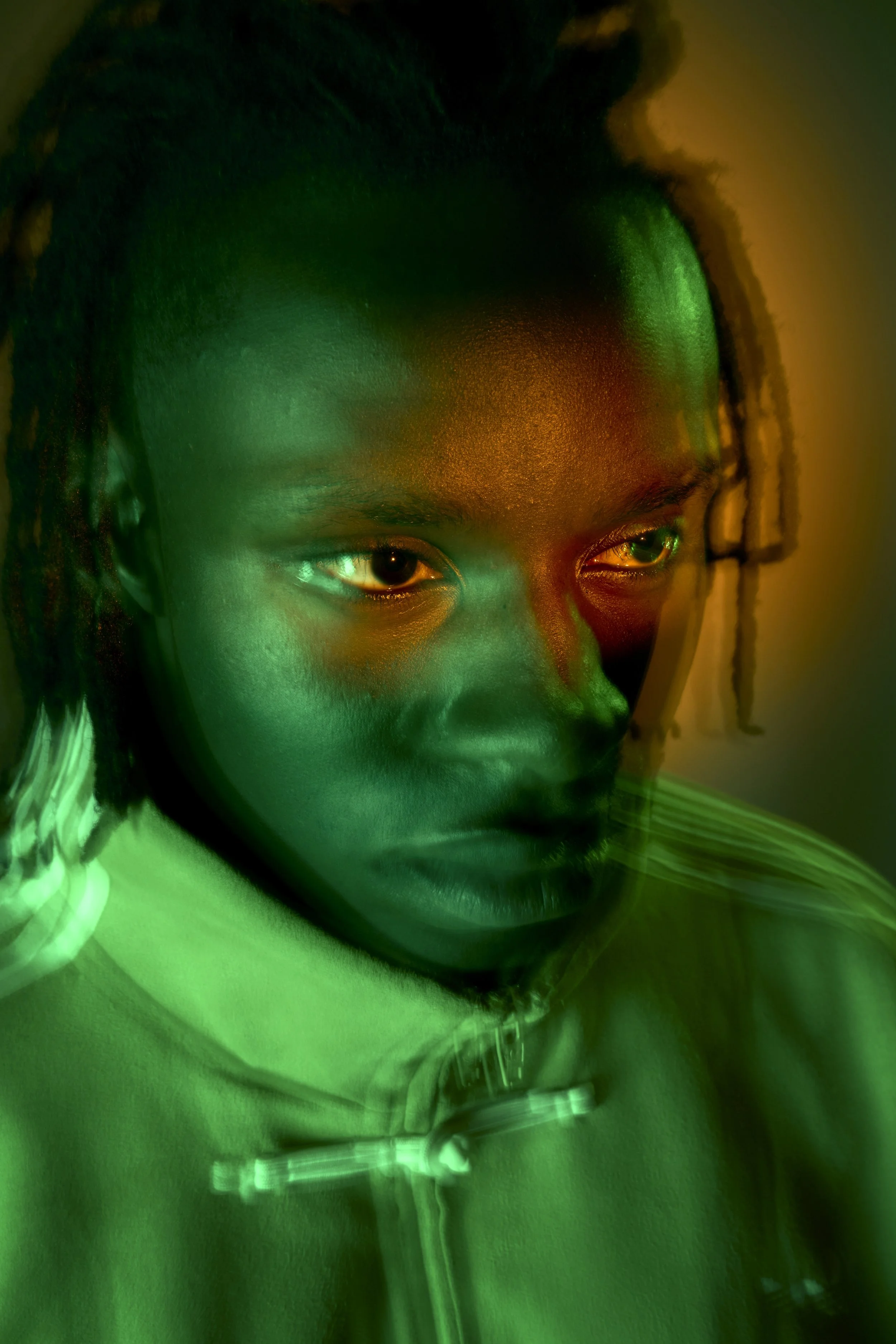 A young Black man with short dreadlocks, shot close and from above, stares into the distance. The lighting is green, blurry and dreamlike, only his eyes are crisp and sharp.