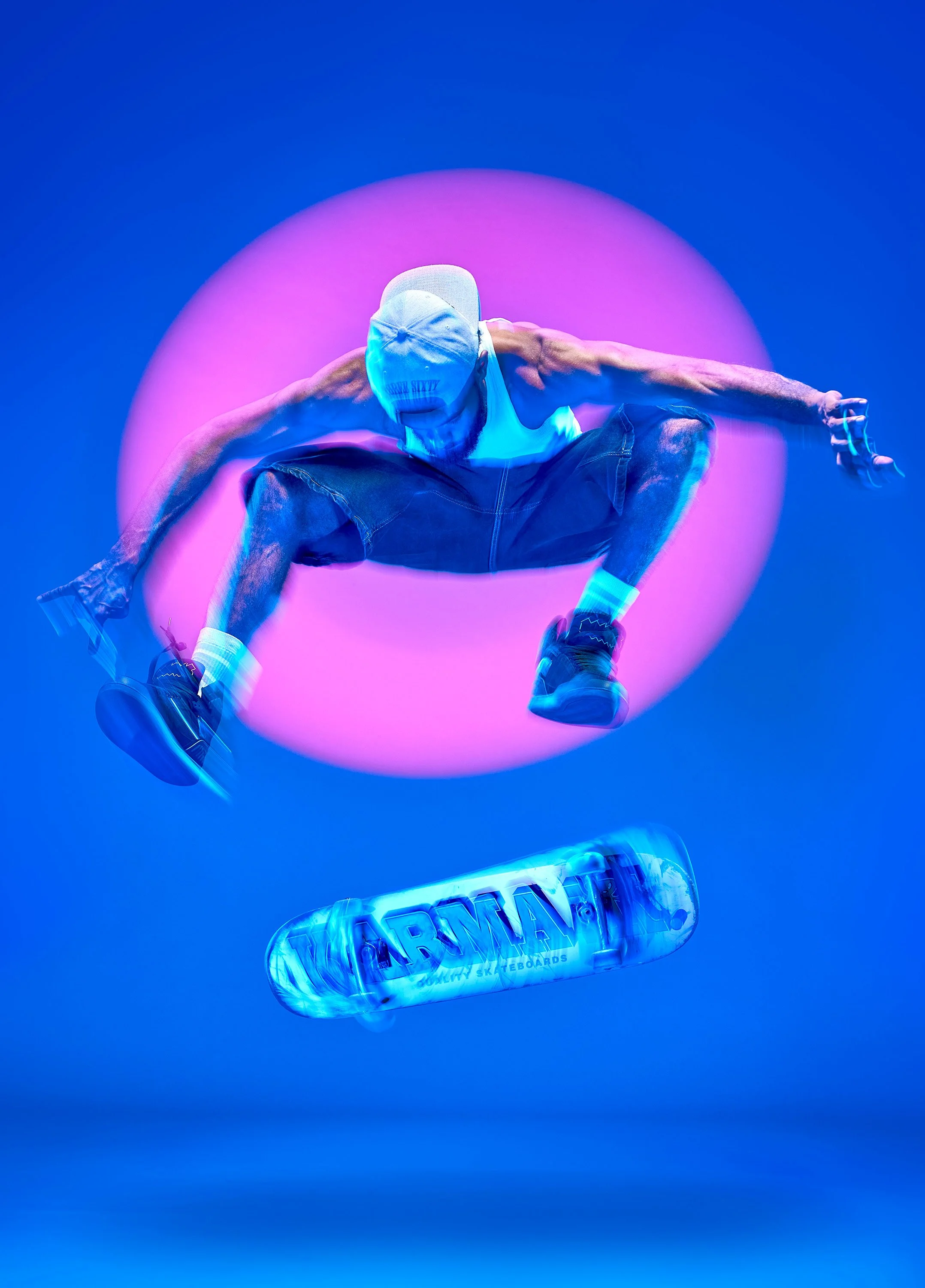 A Black male skateboarder performs a heelflip in a studio. He is frozen by flash but the board and his extremities are motion blurred, and there is a large pink circle of light projected on the wall behind him.