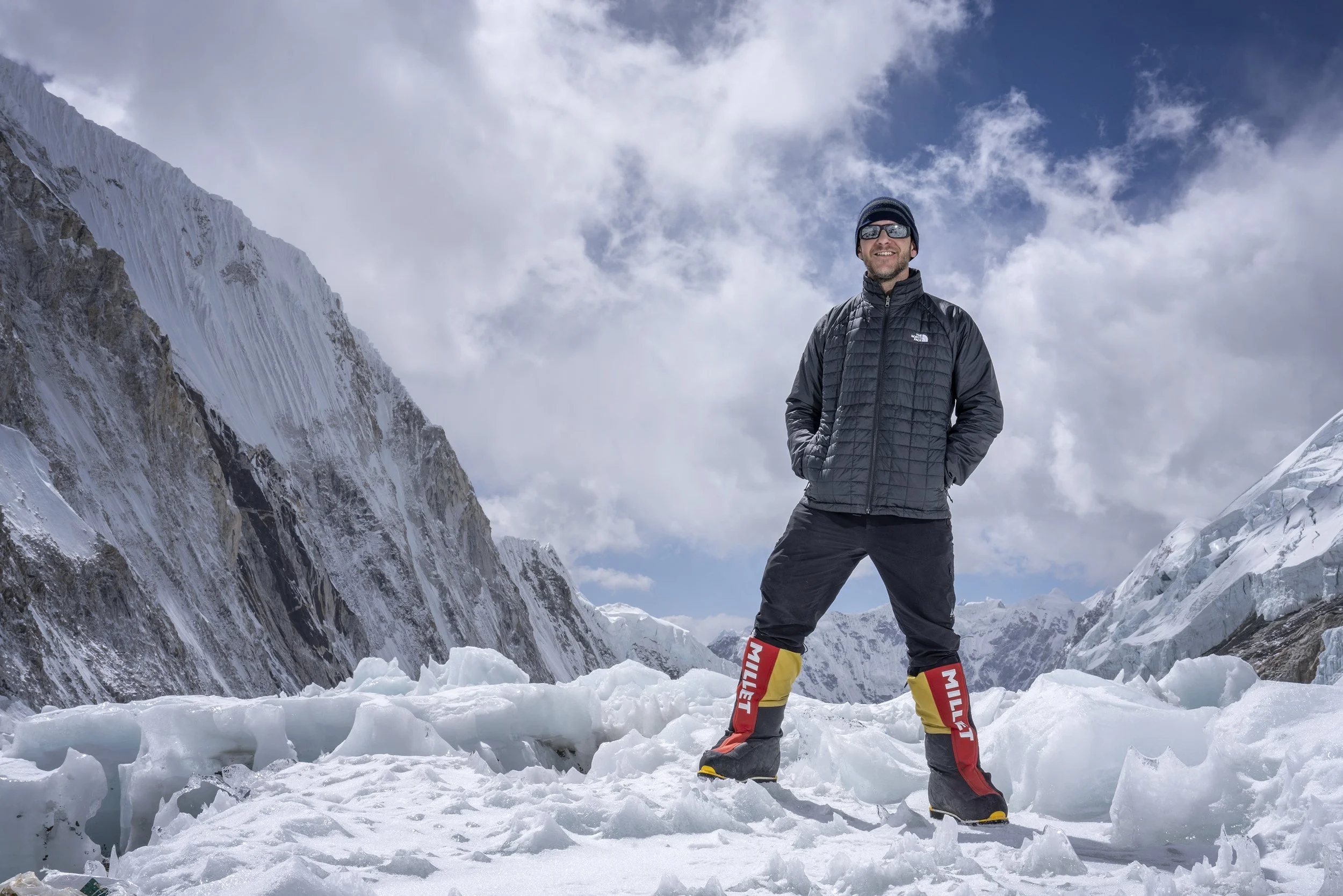 Photographer Steve Bright stands in th snow at Camp Two on Everest, wearing mountaineering boots, sunglasses and a down jacket