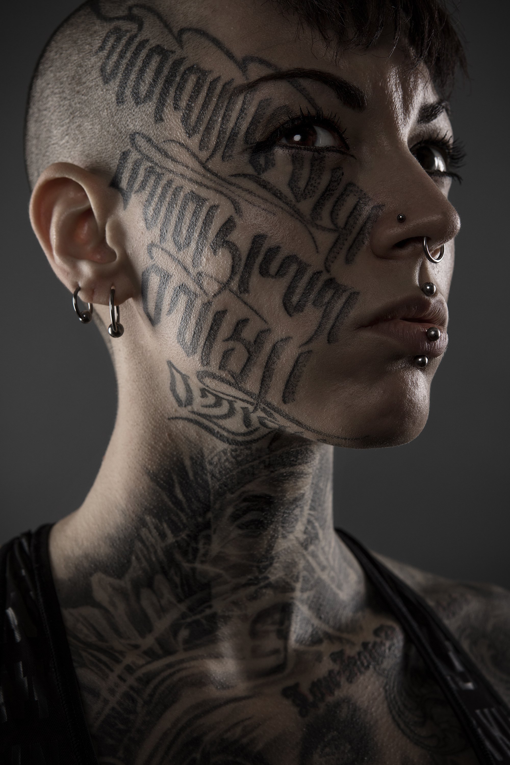 A young woman with a shaved head and elaborate text tattooed across her face, lit moodily from the sides. commercial portrait photography by Steve Bright