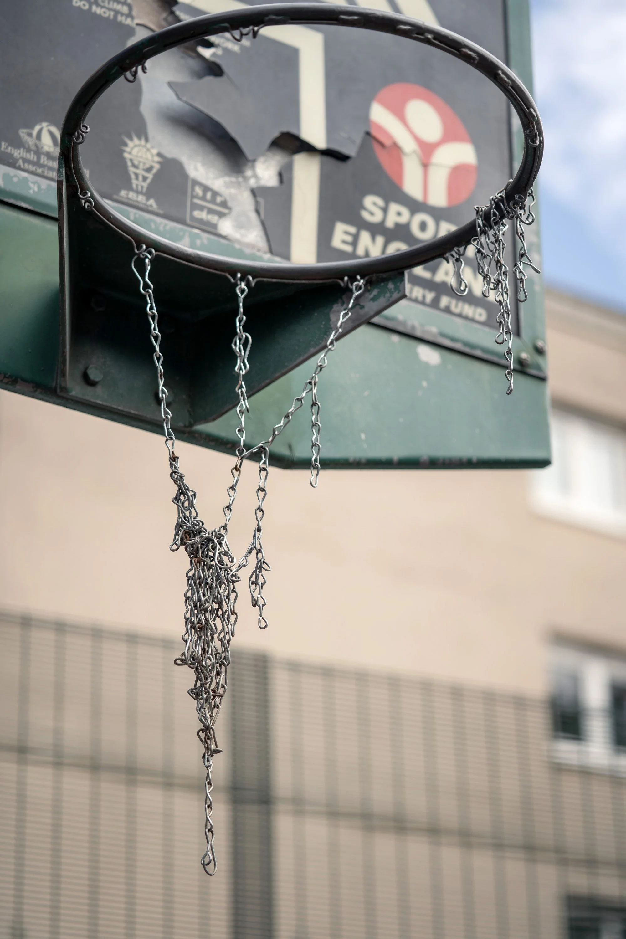 Broken basketball hoop with hanging chains, on an outdoor basketball backboard that has a torn and damaged surface. Dynamic sports portrait photography by Steve Bright