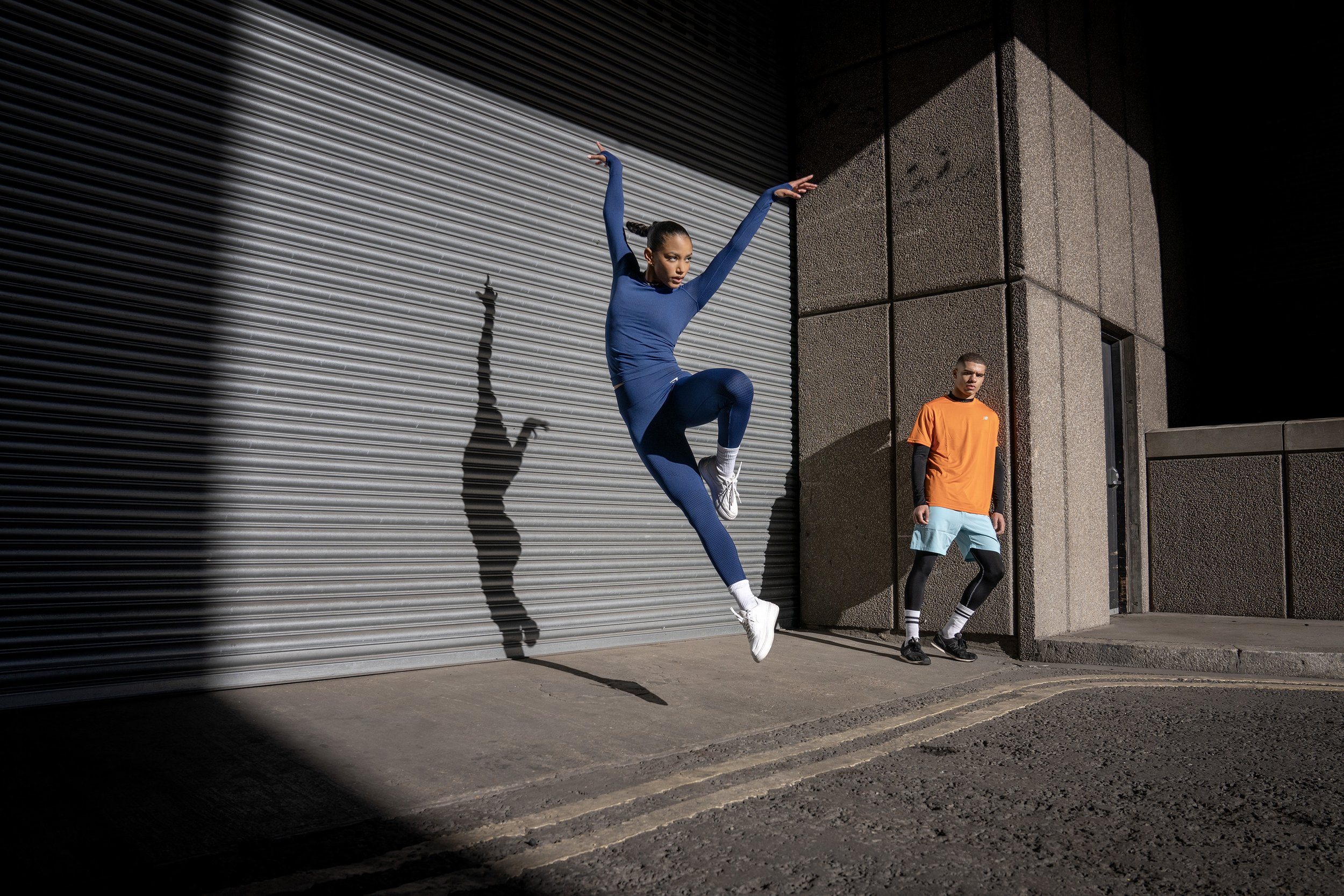 A young man squats in the foreground of a brutalist urban scene, while behind him a young woman leaps gracefully into the air, her shadow cast agains the wall by the harsh sunlight. dynamic sports photography by Steve Bright