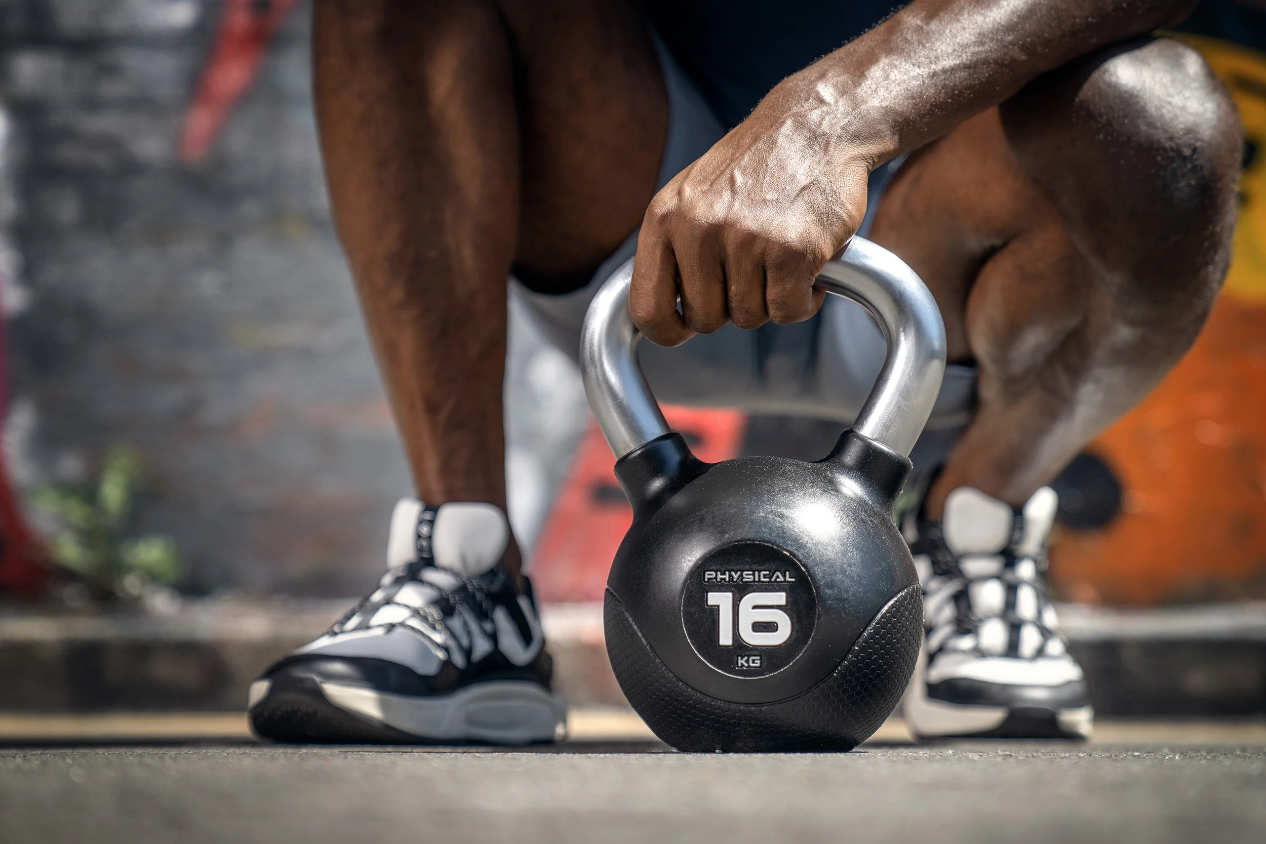 A man squats down by a 16kg kettlebell, wearing black and white sneakers, and grips the handle of the kettlebell. Dynamic sports portrait photography by Steve Bright