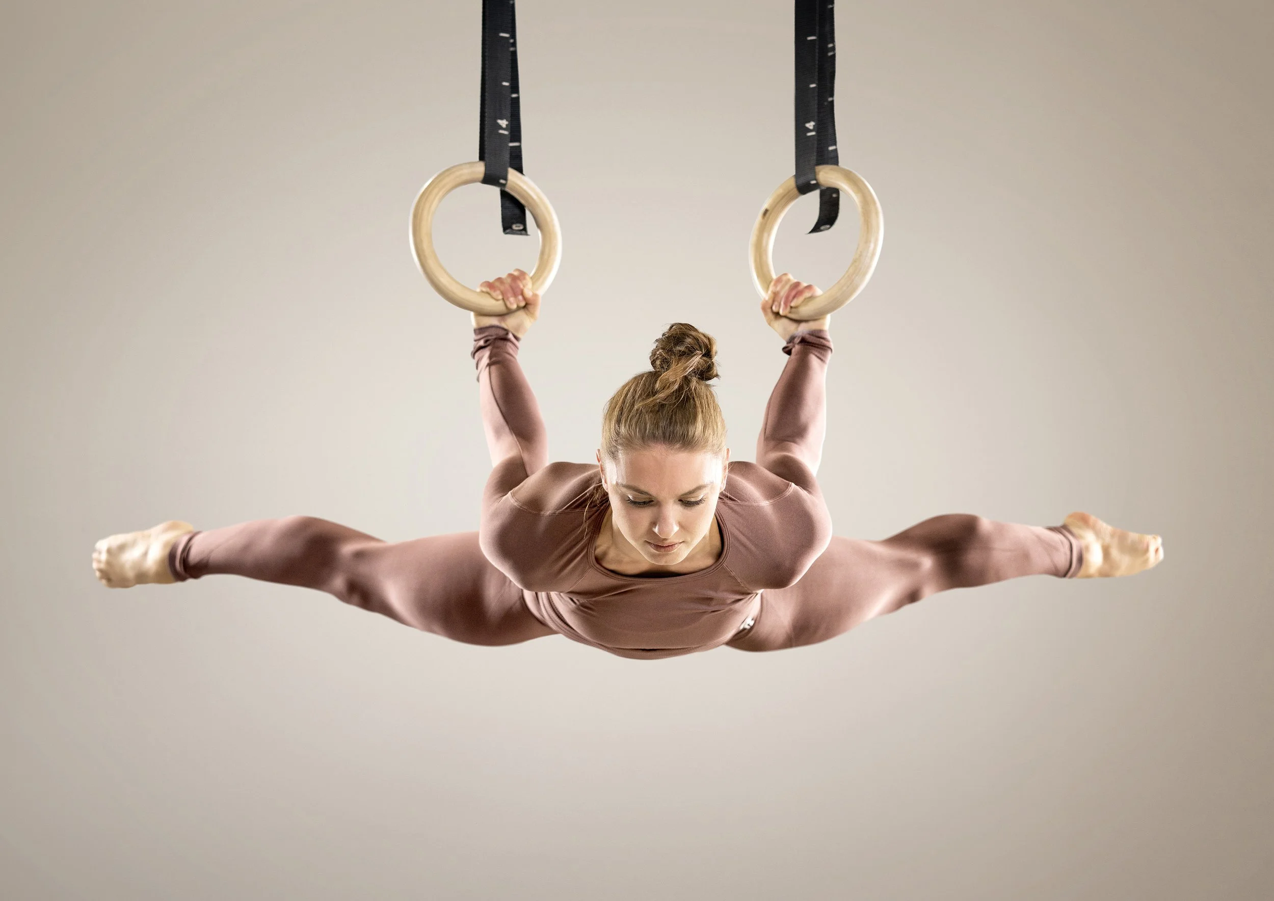 A female gymnast performing a split move on gymnastic rings, wearing a brown athletic outfit, with arms extended. dynamic sports  portrait photography by Steve Bright