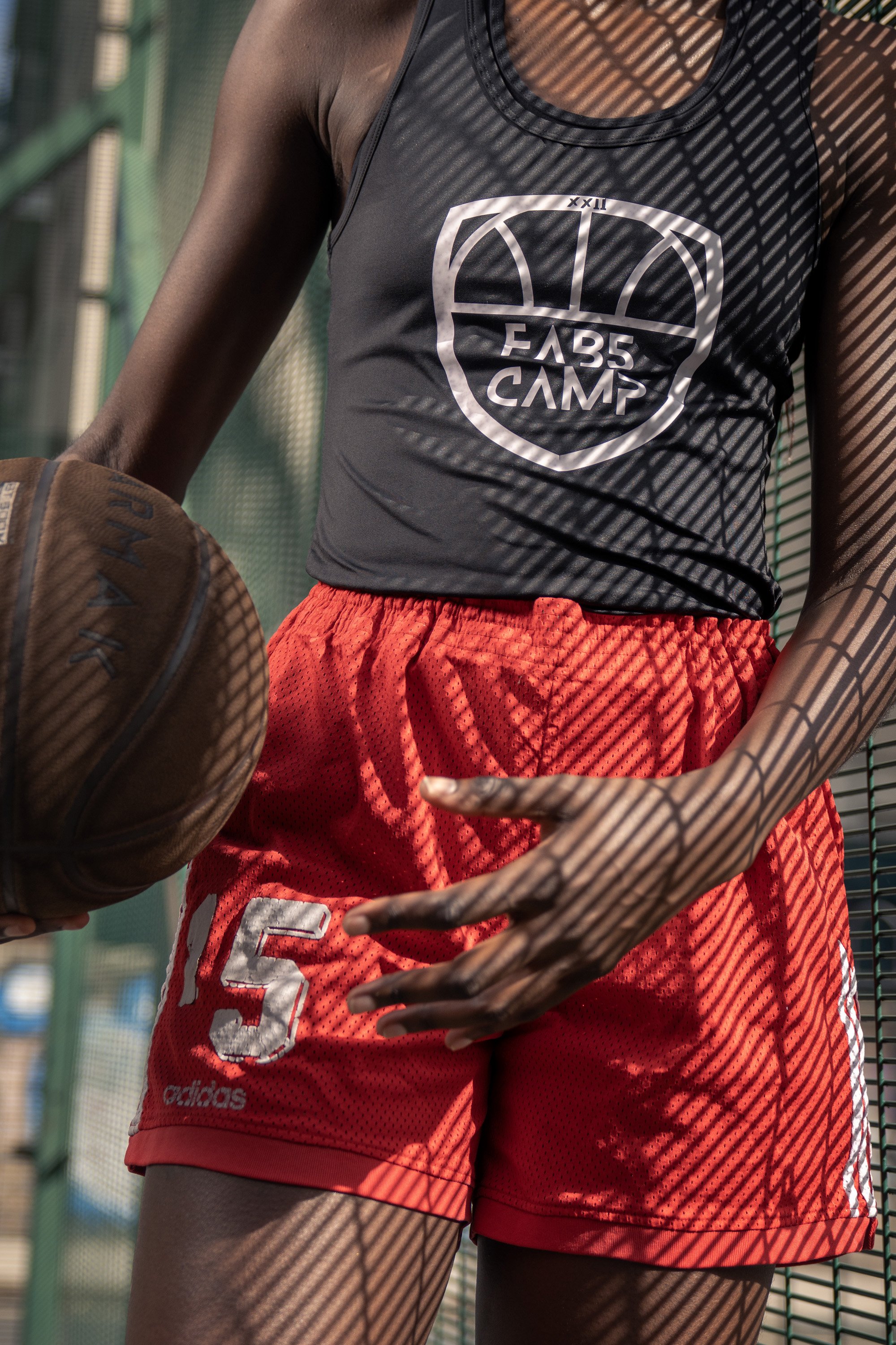 Close-up of a young woman standing on an outdoor basketball court, leaning against a wire fence with the shadows of the fence cast on her face and body, holding a basketball, dynamic sports  portrait photography by Steve Bright