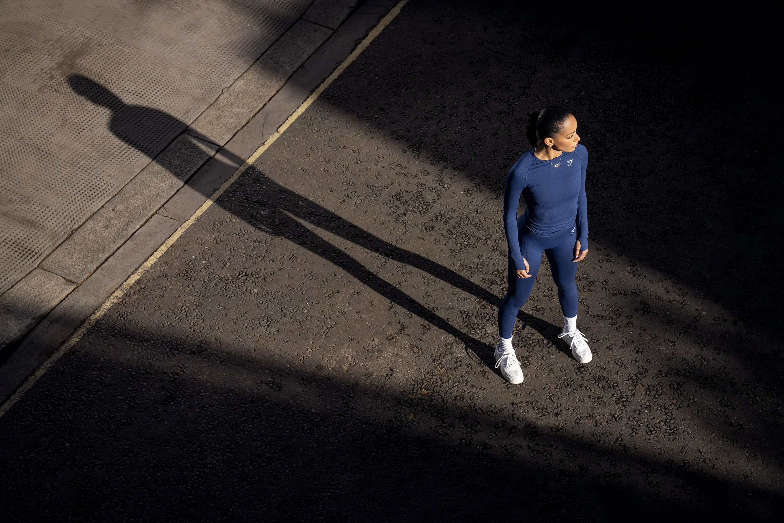 A young woman in blue fitness clothing is seen from above, standing in a beam of sunlight on an urban road, her long shadow cast behind her. dynamic sports portrait photography by Steve Bright