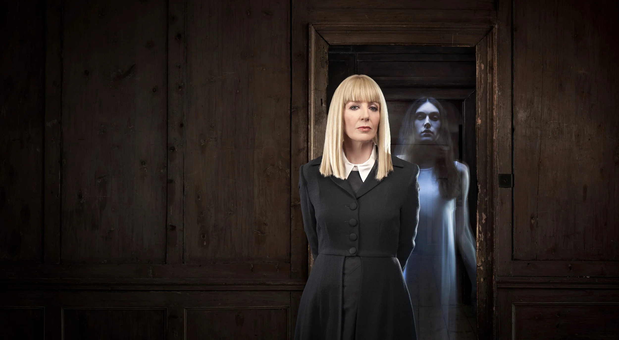 Publicity image for tv show Most Haunted. Presenter Yvette Fielding stands in front of a doorway wearing a black coat, while a ghostly figure stands behind him in the doorway. portrait photography by Steve Bright, London