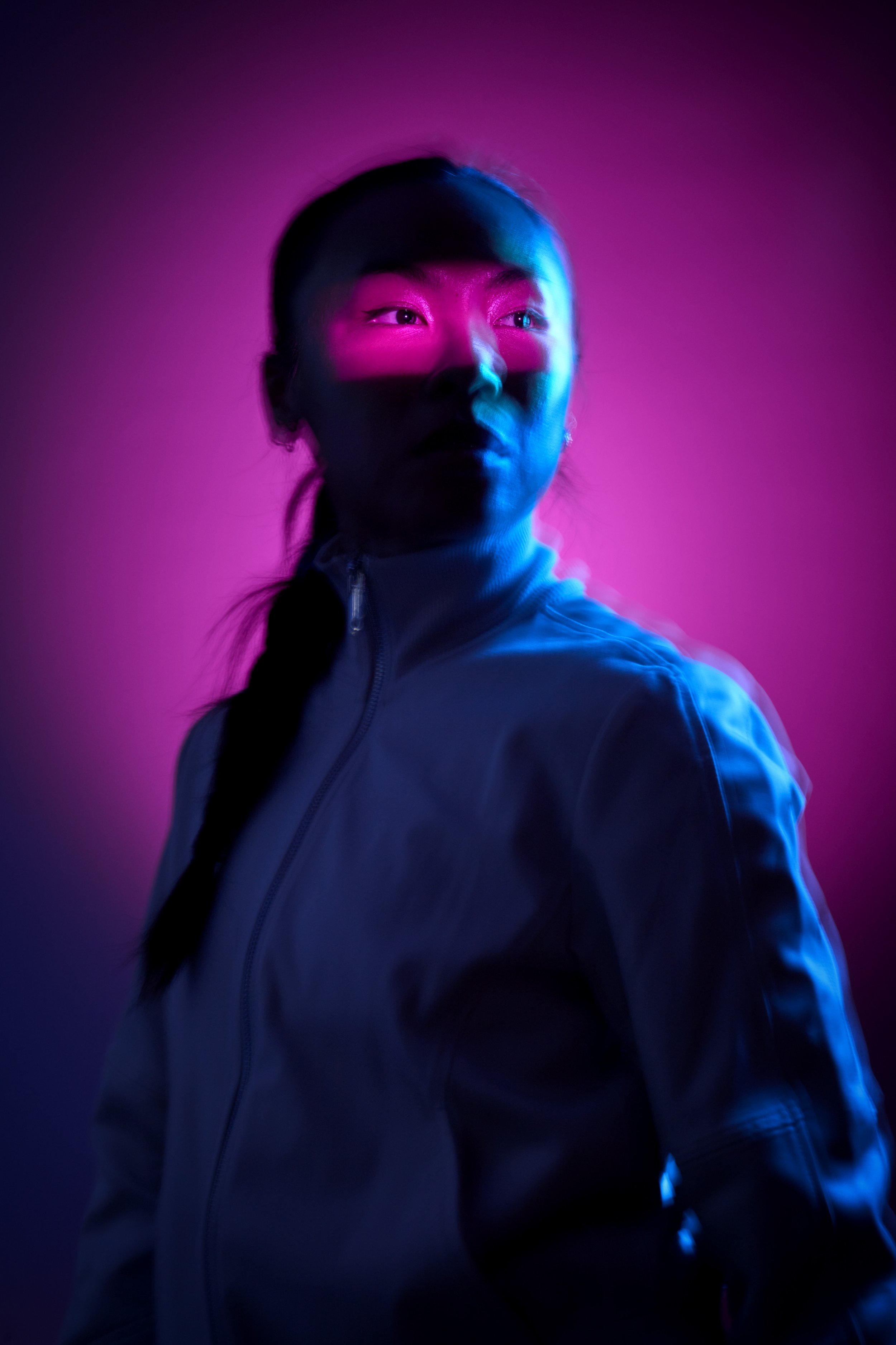 A young Asian woman is almost silhouetted against a moody pink backgrond, her outline motion blurred and indistinct. The only part of her that is lit is her eyes, crisp and sharp in a strip of pink light projected onto her face.