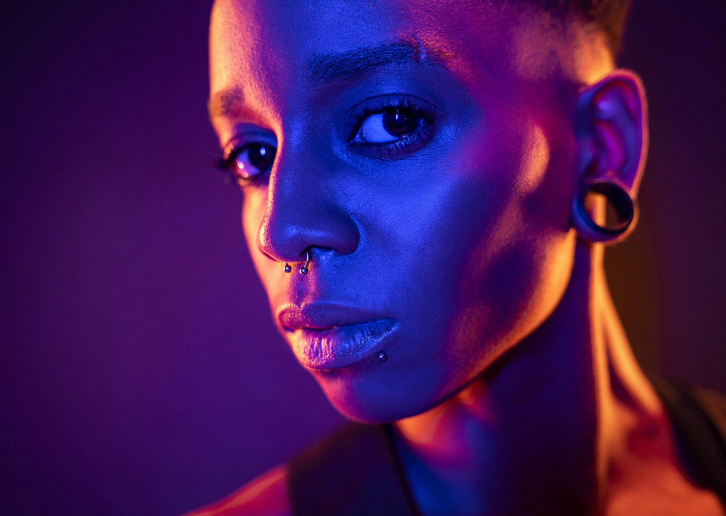 A cool young Black woman with short hair and piercings lit with neon coloured lighting, commercial portrait photography by Steve Bright