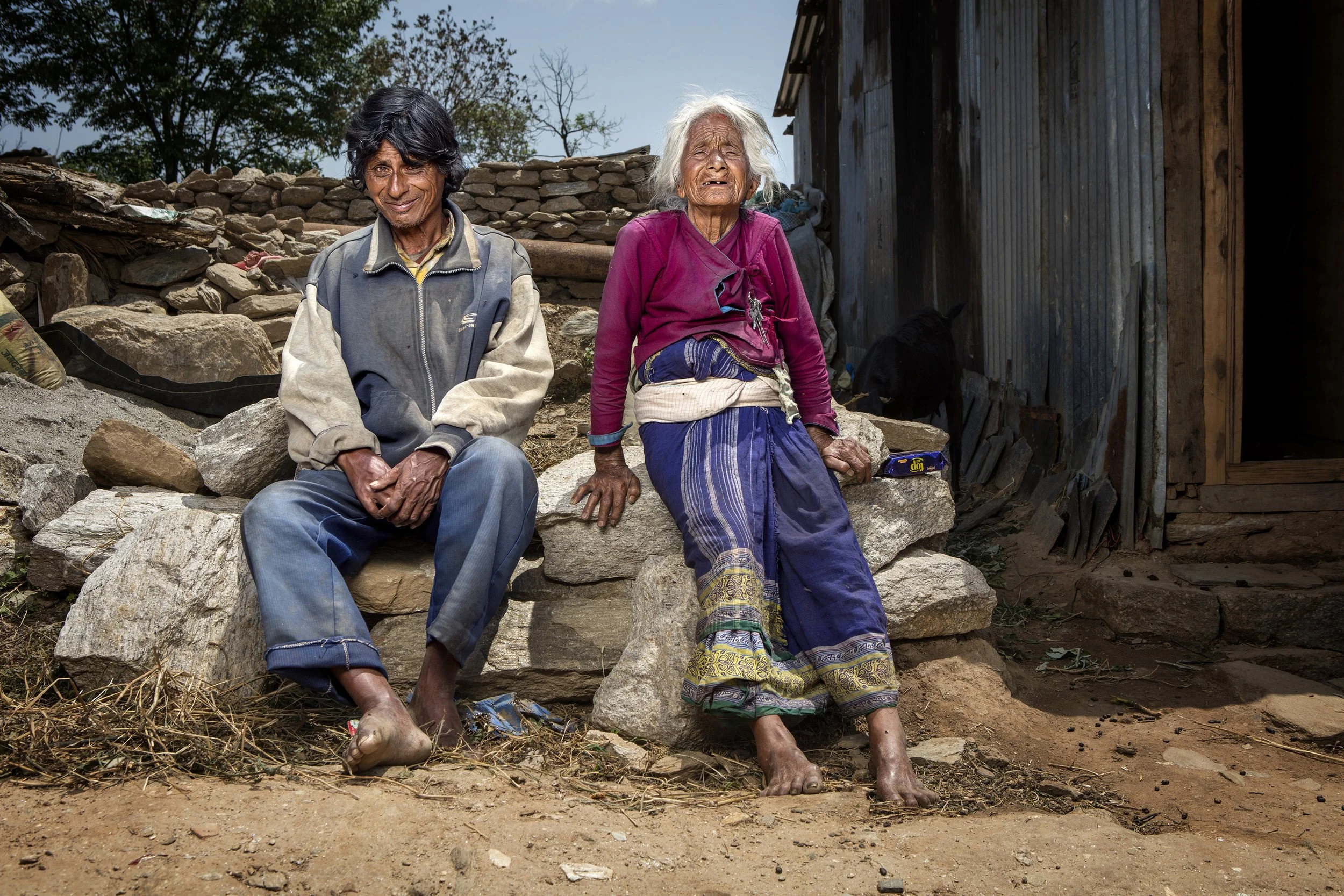 An elderly nepalese lady and her disabled adult son sit on a wall outsode their metal shack, after their house was destryed by the 2015 eathquake. commercial portrait photography by Steve Bright