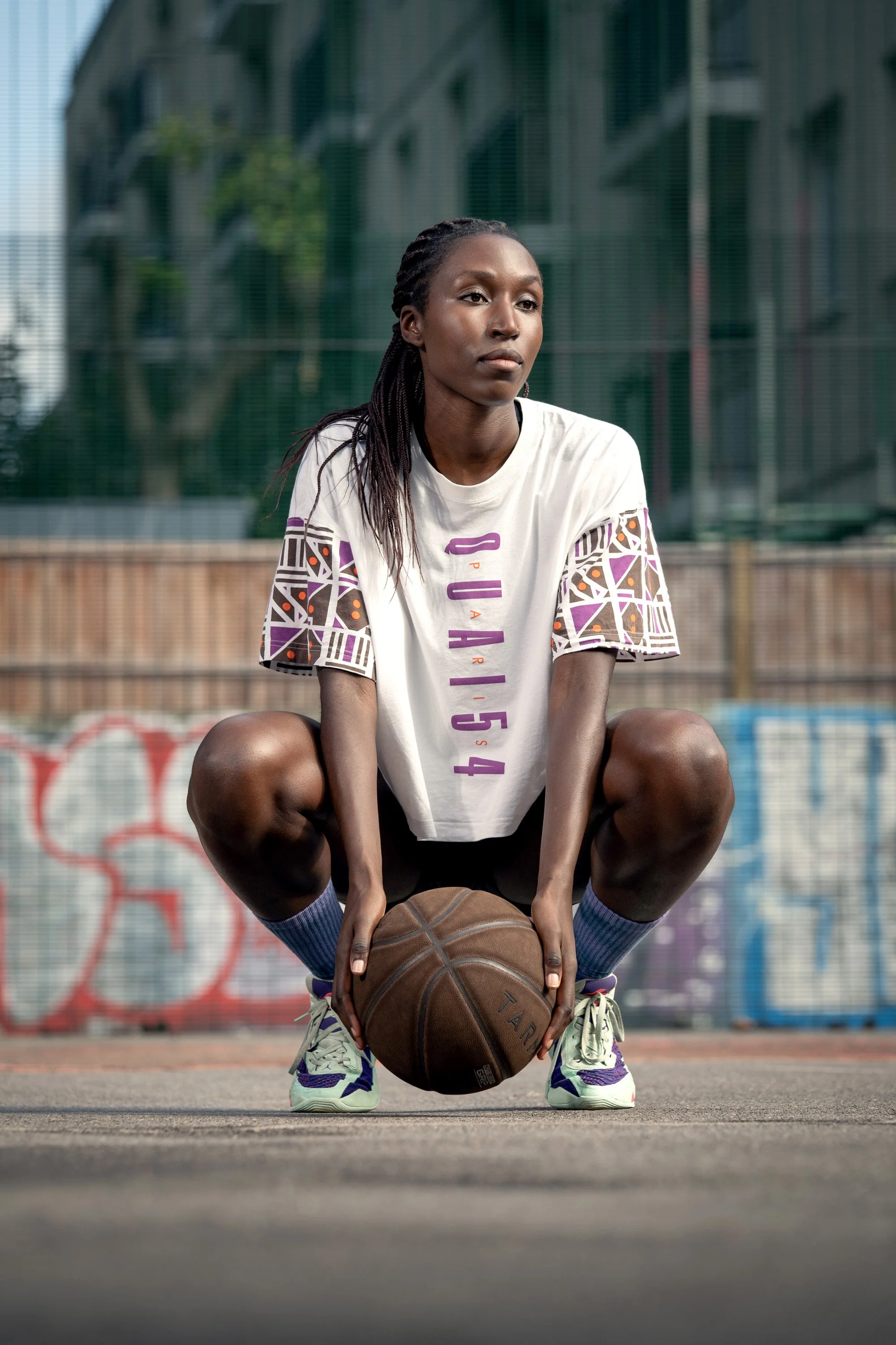 A woman on a basketball court holding a basketball, in an athletic stance, wearing a white t-shirt and purple sneakers. Dynamic sports portrait photography by Steve Bright
