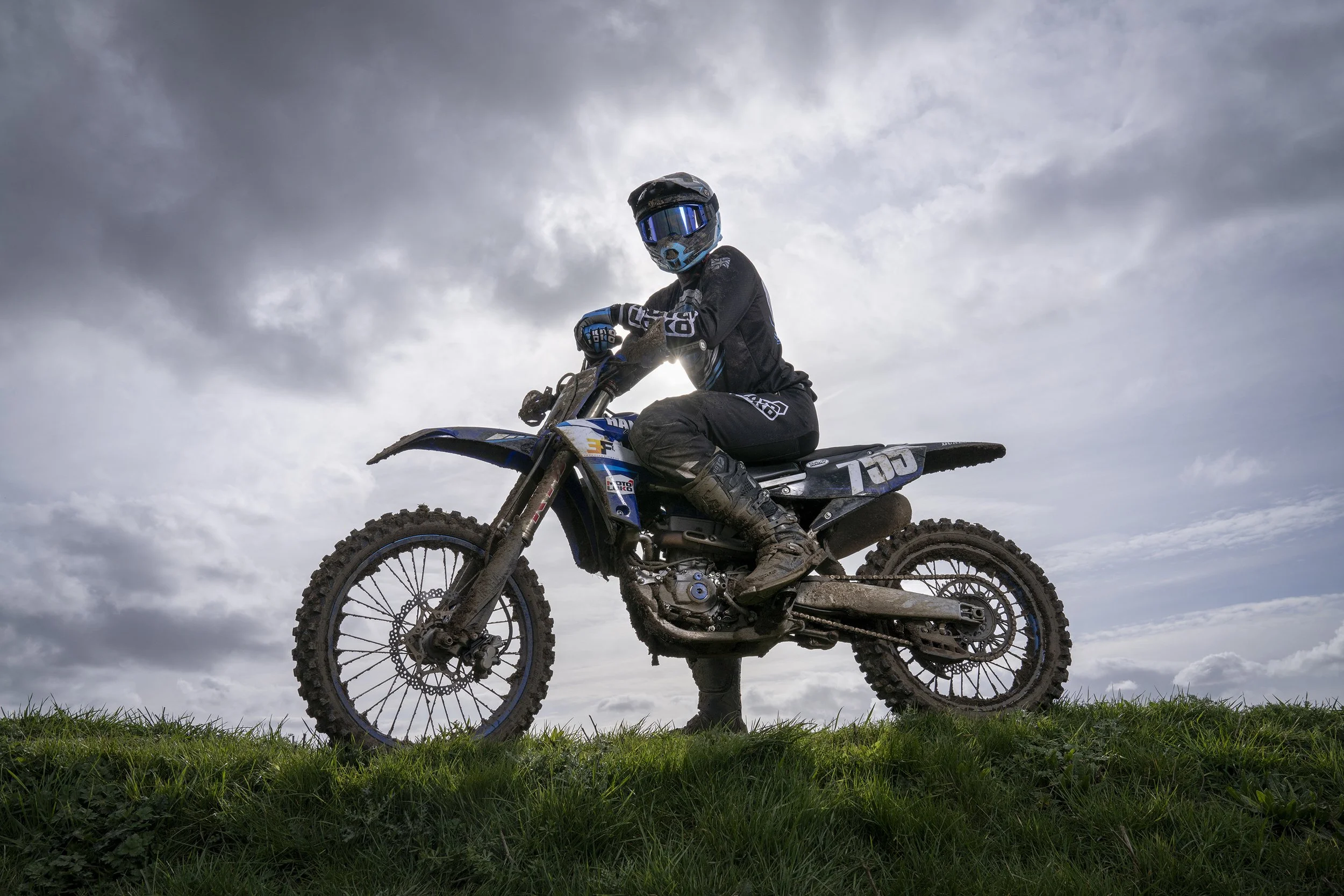 A person in motocross gear sitting on a dirt bike on a grassy hill with a cloudy sky in the background. dynamic sports portrait photography by Steve Bright
