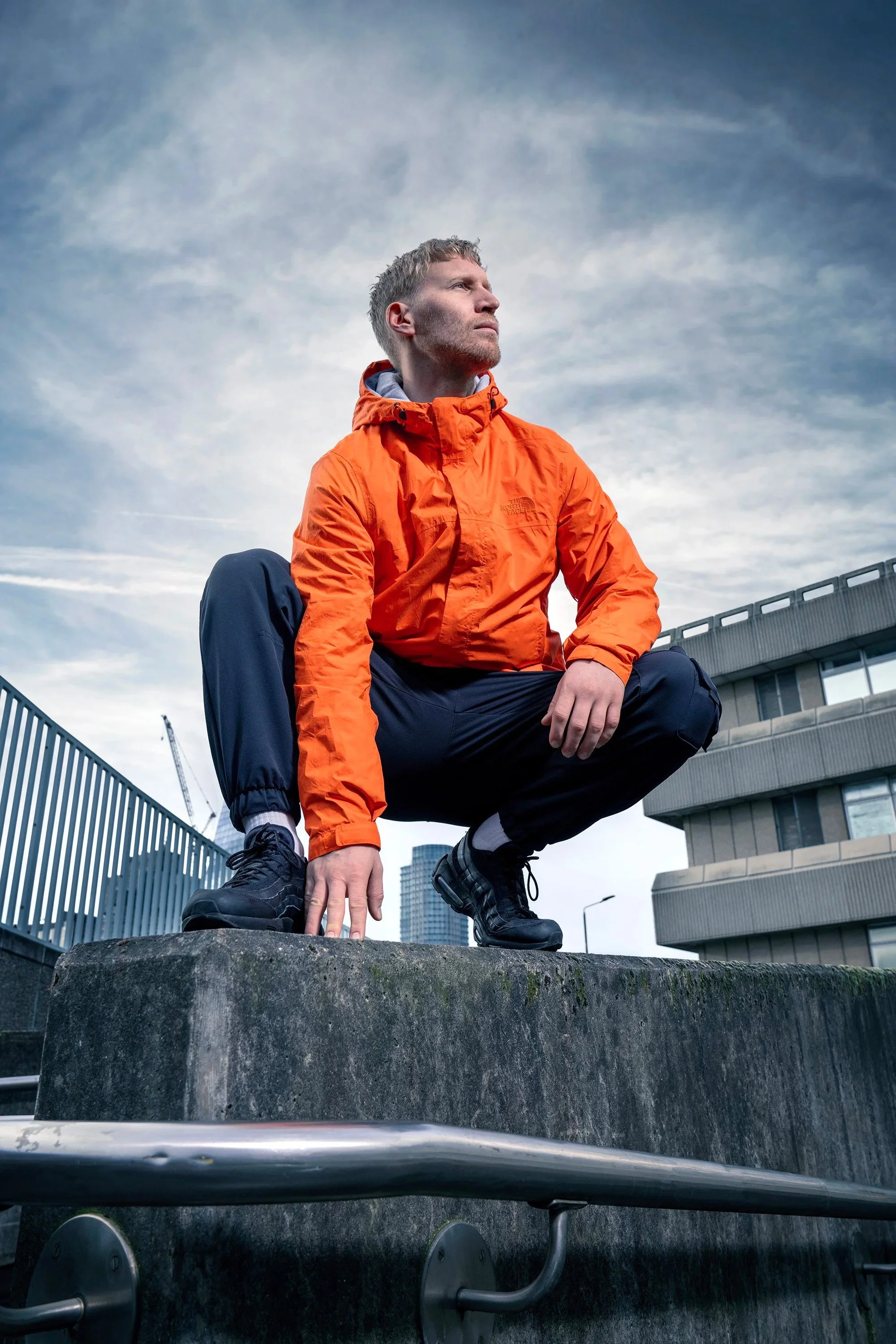 A man crouching on a concrete ledge outdoors, wearing an orange jacket, with city buildings and a cloudy sky in the background. dynamic sports  portrait photography by Steve Bright