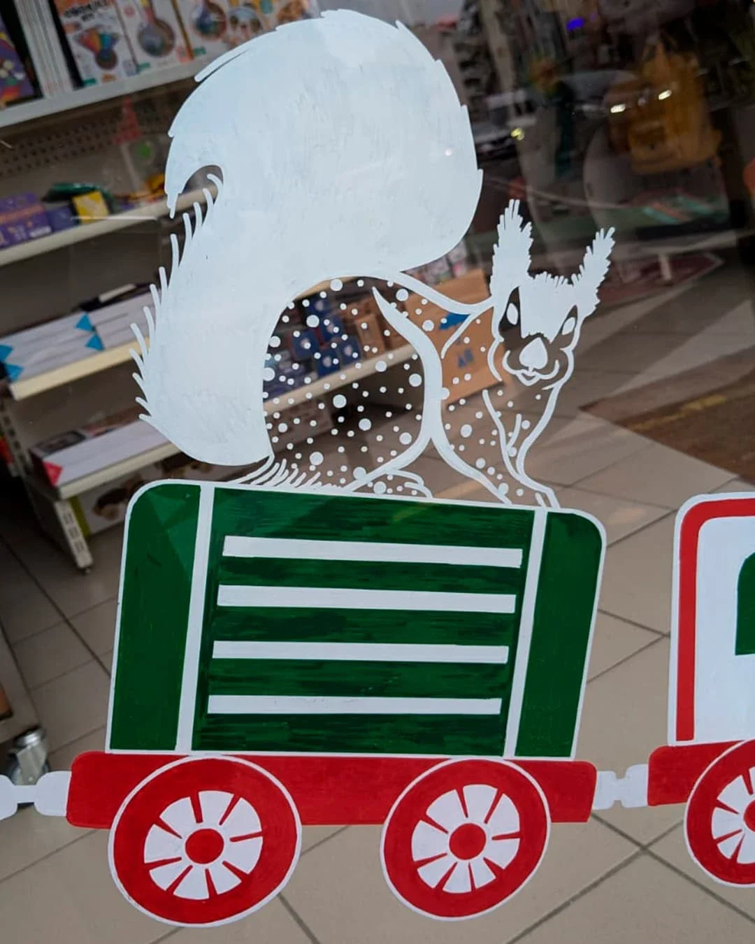 Peinture sur vitrine de Noël sur-mesure pour une boutique de jouets à Houilles (78), avec écureuils et vieux train d'autrefois, coloris blanc, rouge et verts