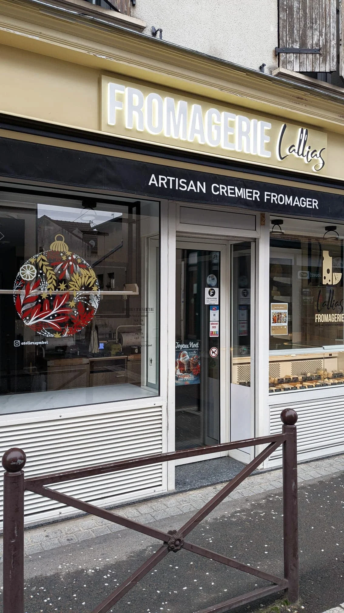 Façade de la fromagerie Lallias à Houilles décorée d'une peinture sur vitrine de Noël réalisée à la main avec une grande boule de Noël ornée de végétaux et d'une tranche d'agrume.