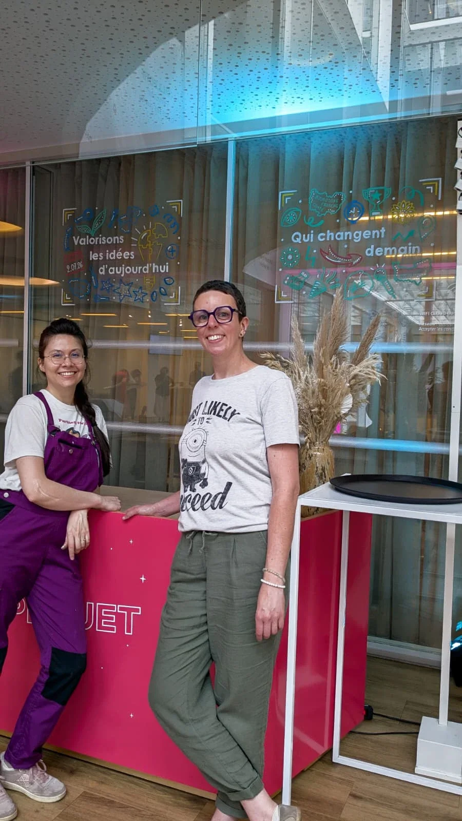 Peinture sur vitrine événementielle pour l'Apec dans le cadre des Trophée de l'Innovation 2026. Prestation dans les locaux de l'événement à Paris. Sandrine Daniel et sa collaboratrice Camille Fauquet devant 2 visuels peints.