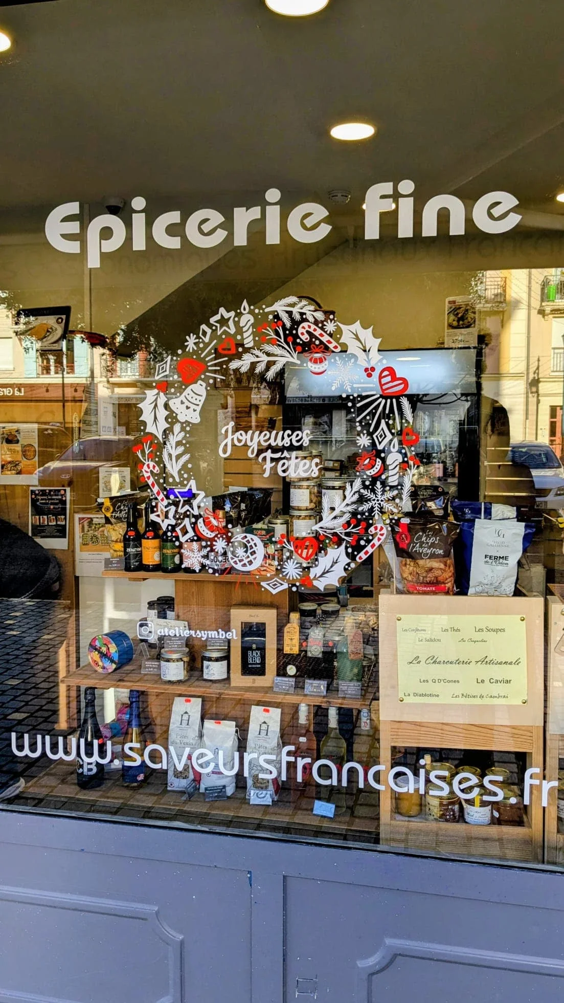 Vue rapprochée de la couronne Doodle peinte à la main sur la vitrine de Noël de l'épicerie Saveur Française à Vauréal (95), composée de houx, cloches, étoiles, sucre d'orge et branches de pin