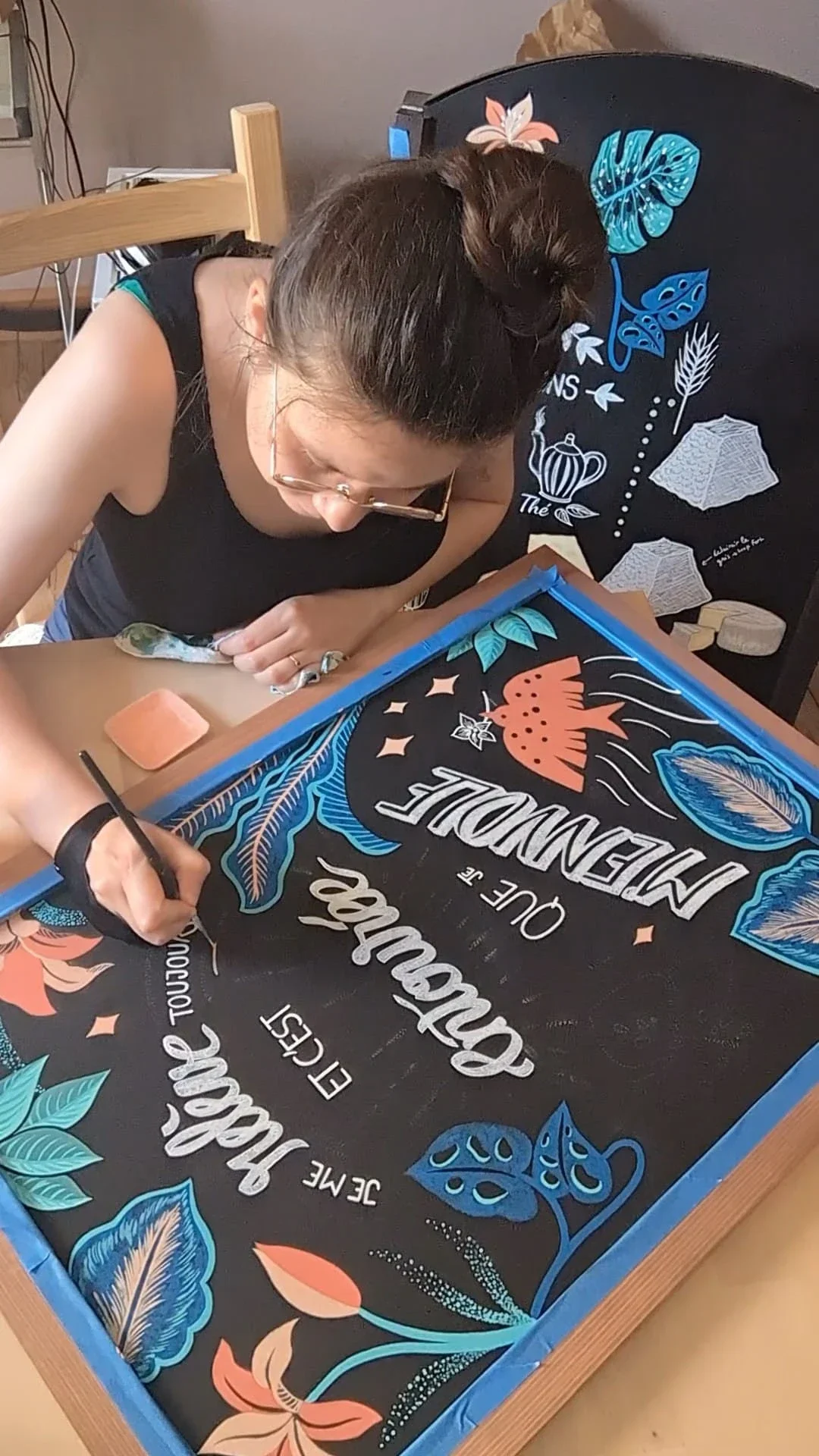 Sandrine Daniel ajoute des lignes ornementales peintes à la main autour du mot « toujours » sur une ardoise décorative