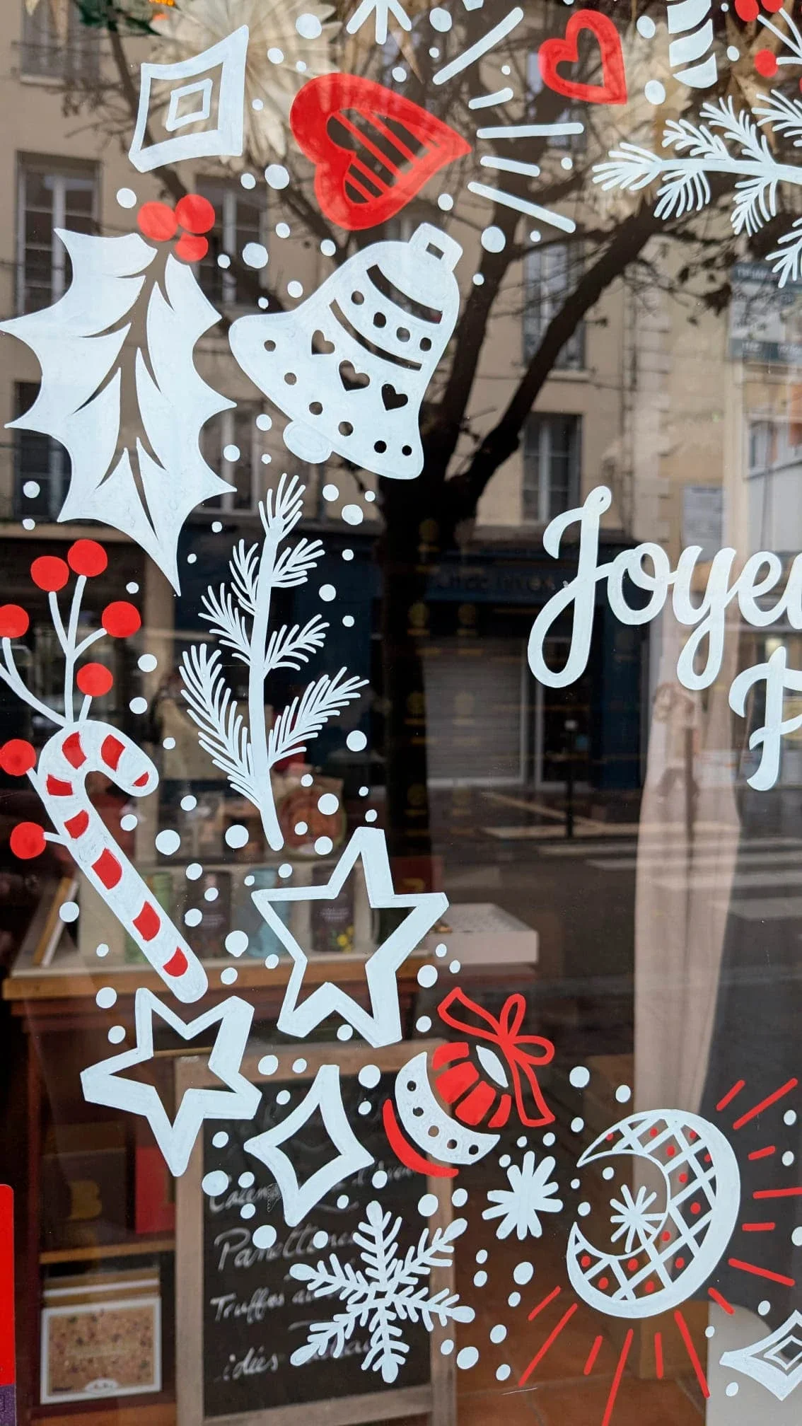 Peinture sur vitrine représentant une couronne de Noël peinte à la main. Détail sur le motif composé de houx, cloches, étoiles, baies et branches de pin. Réalisée pour l'épicerie fine Le Rameau d'Olivier à Saint-Germain-en-Laye (78)