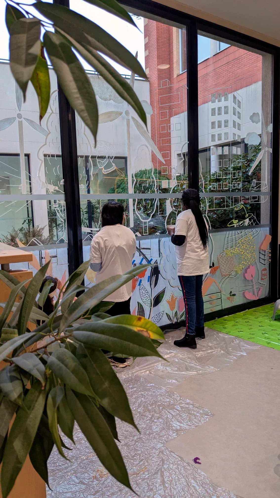 Fresque participative sur les vitrines du patio de la médiathèque des Mureaux, réalisée lors des Nuits de la lecture 2026. Thème "Villes et campagnes". Deux jeunes filles peignent sur la fresque. Peinture acrylique.