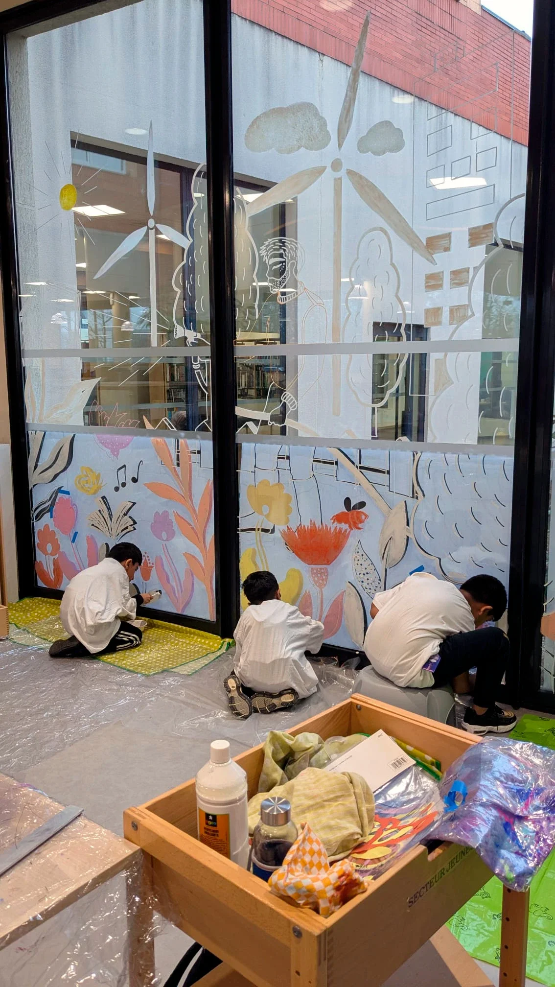 Fresque participative sur les vitrines du patio de la médiathèque des Mureaux, réalisée lors des Nuits de la lecture 2026. Thème "Villes et campagnes". 3 garçons peignent sur la fresque.