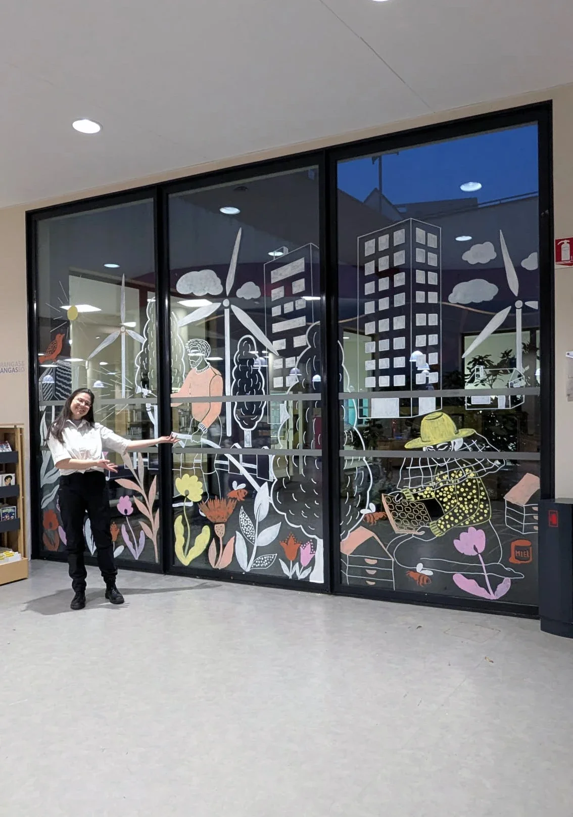 Peinture sur vitrine. Fresque participative sur les vitres du patio de la médiathèque des Mureaux (78). Dans le cadre des Nuits de la lecture 2026, thématique "Villes et campagnes". Fleurs, ruches, personnages, éoliennes, arbres. Blanc et couleurs