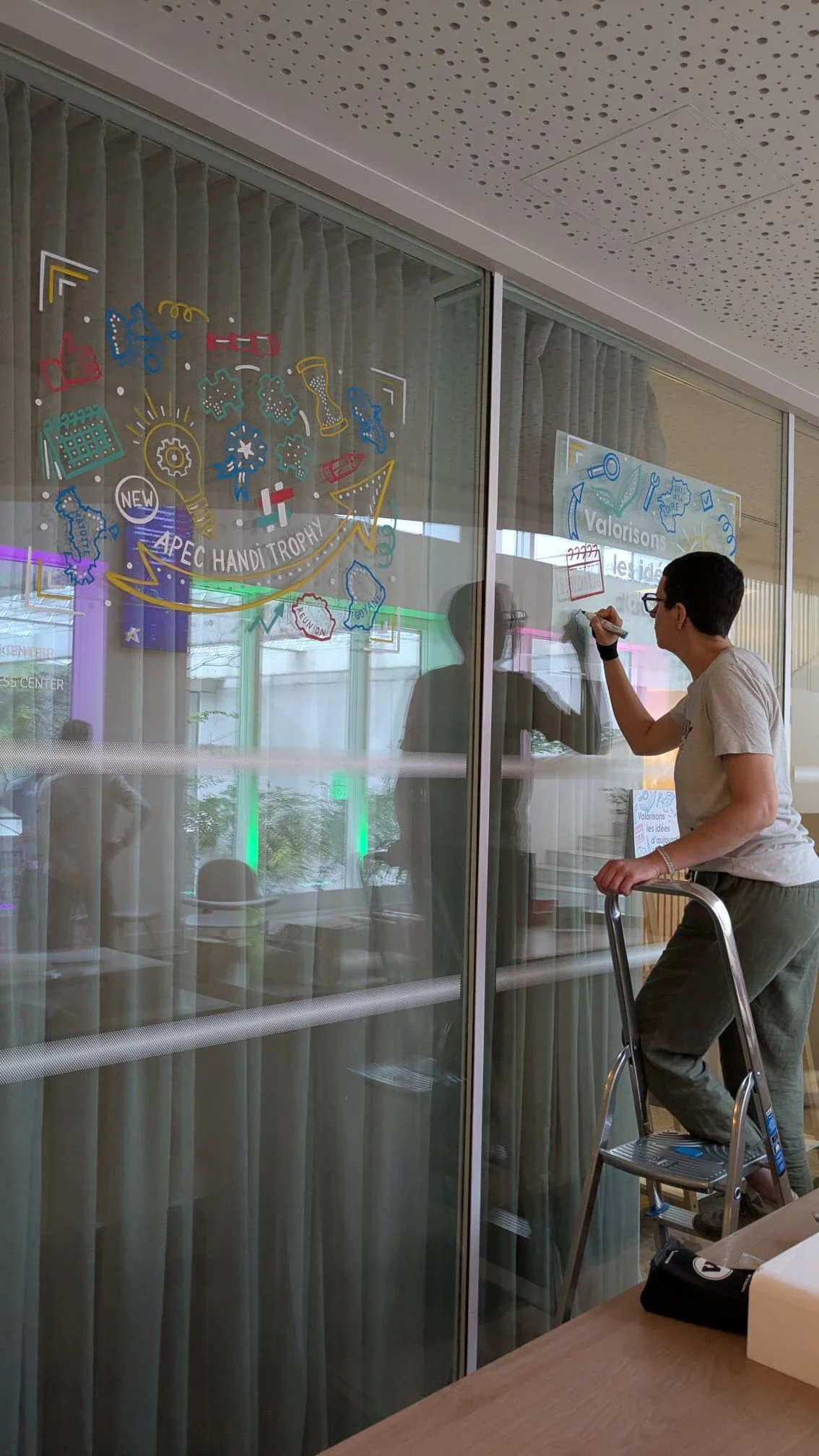 Camille Fauquet peint une vitrine sur son escabeau lors de la réalisation des décors des Trophées de l'innovation de l'Apec