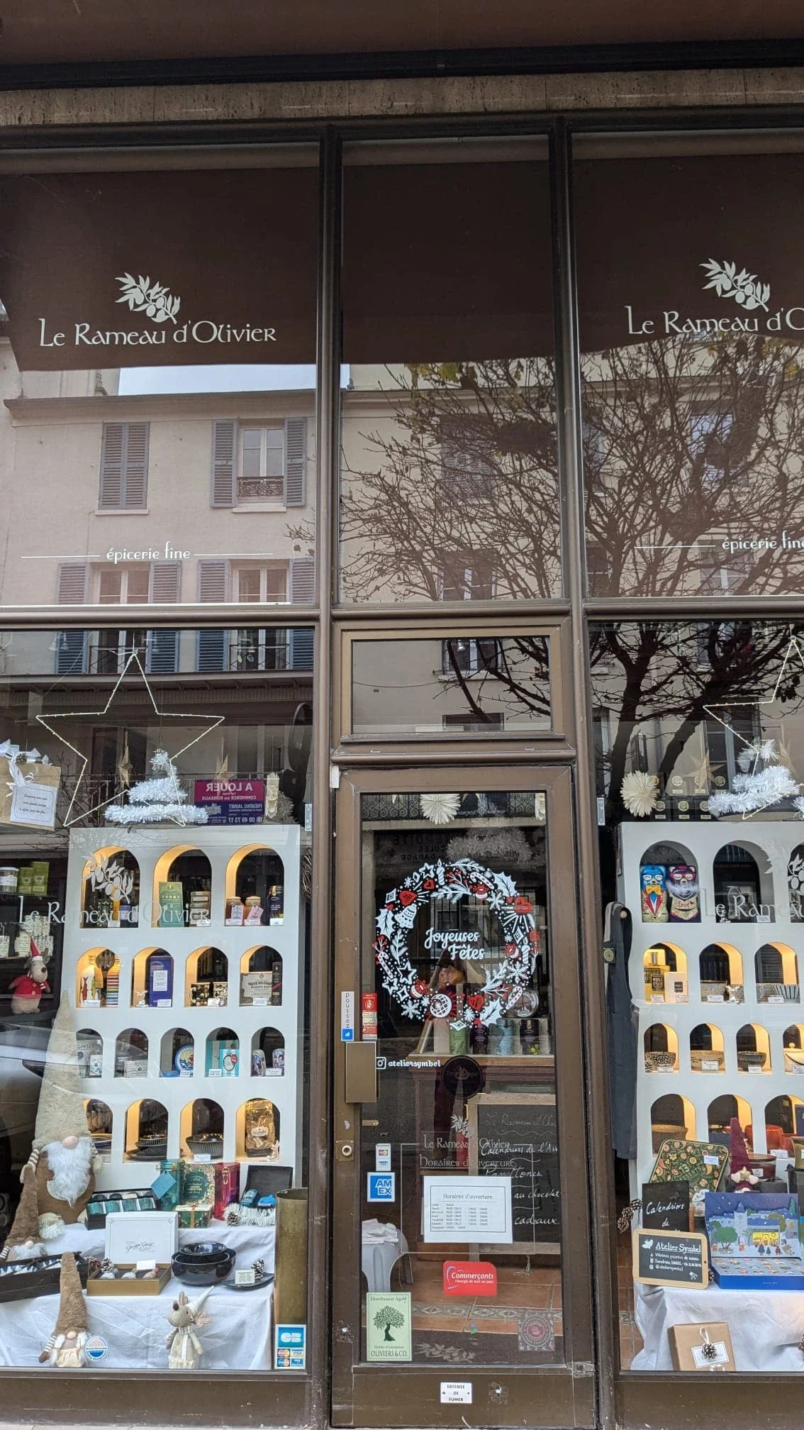 Peinture sur vitrine représentant une couronne de Noël peinte à la main avec houx, cloches, étoiles, baies et branches de pin pour l'épicerie fine Le Rameau d'Olivier à Saint-Germain-en-Laye (78). Plan large sur la devanture de la boutique.