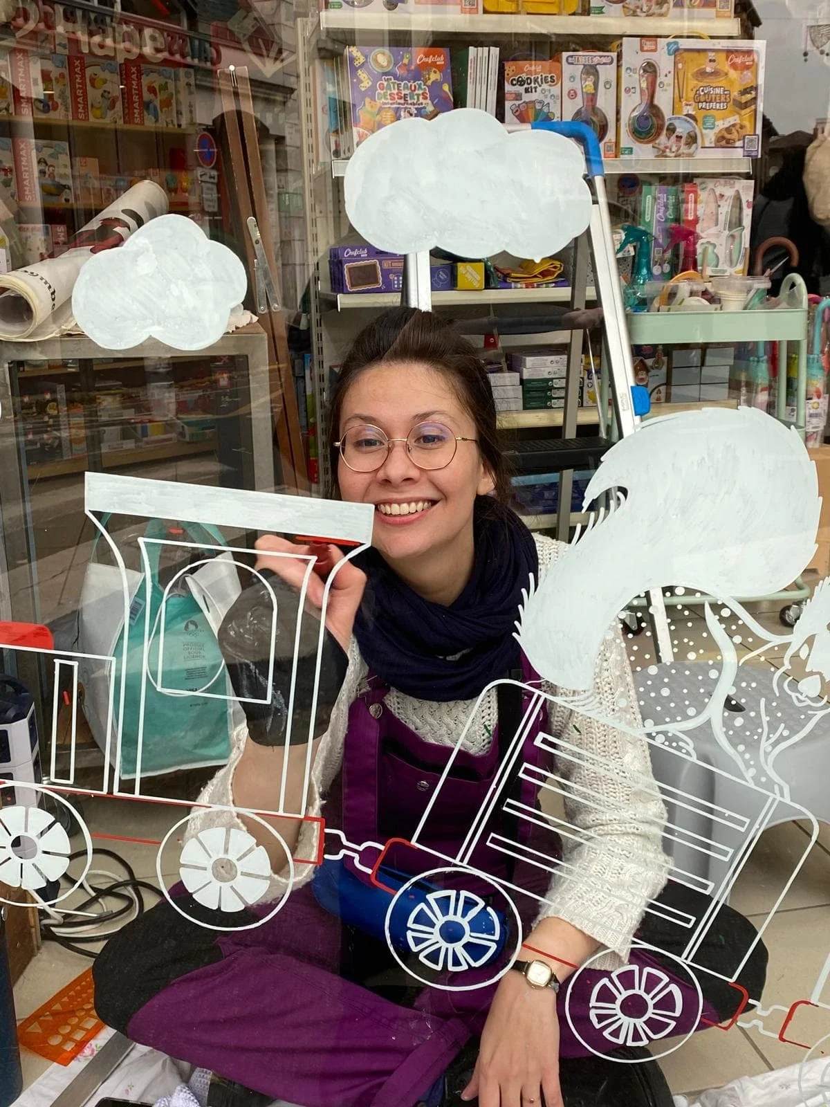 L'illustratrice Sandrine Daniel en train de peindre un train d'autrefois sur la vitrine d'une boutique de jouets à Houilles, hiver 2024