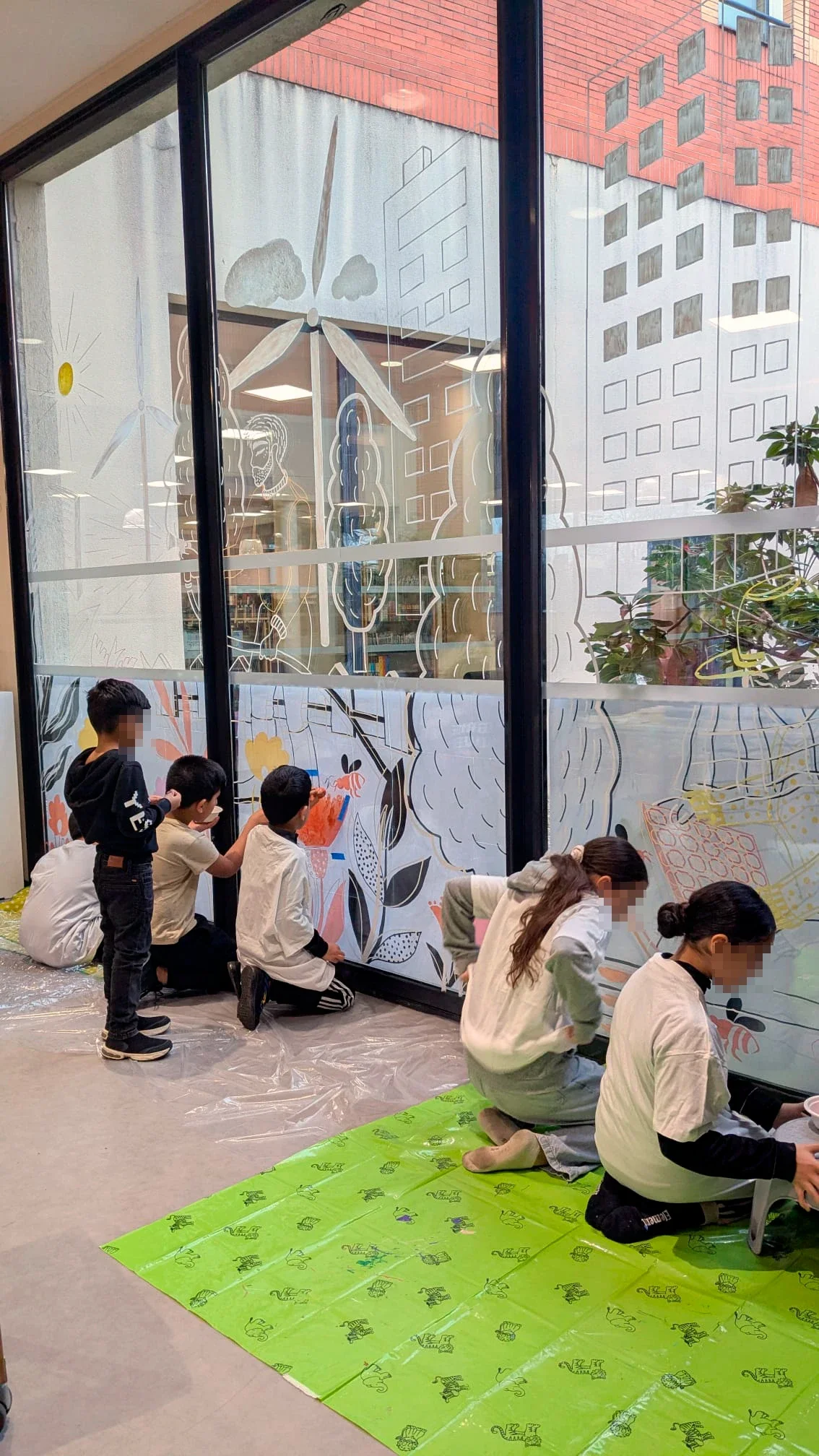 Fresque participative sur les vitrines du patio de la médiathèque des Mureaux, réalisée lors des Nuits de la lecture 2026. Thème "Villes et campagnes". Le public s'empare des différents outils : pinceaux, tampons, pochoirs, marqueurs acryliques Posca