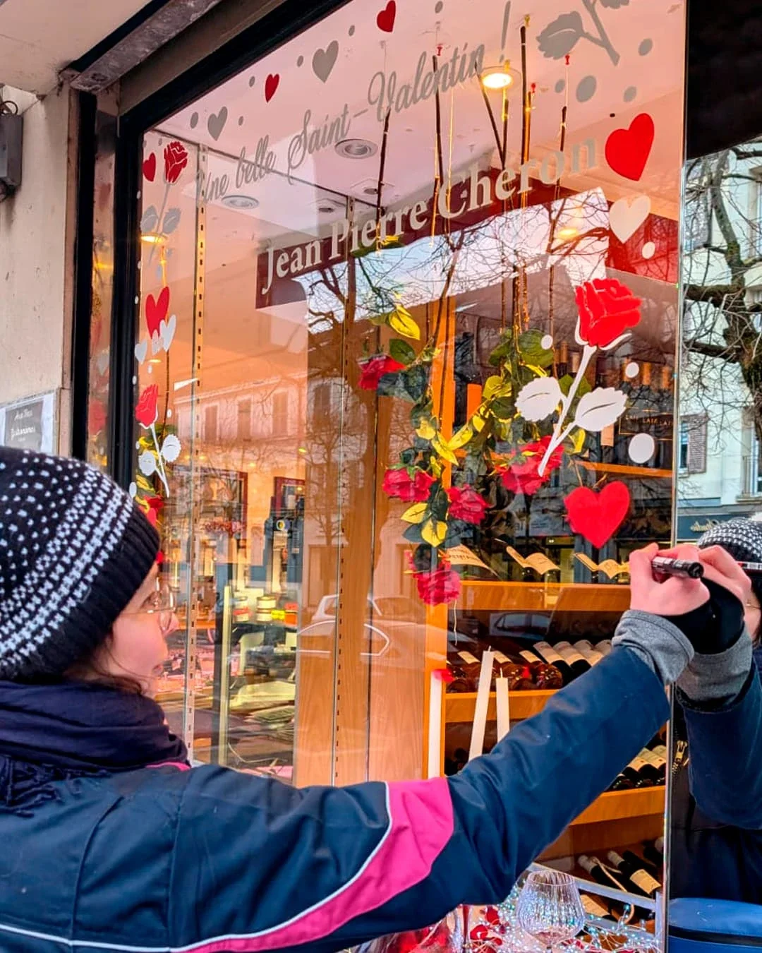 Peinture sur vitrine, thématique Saint-Valentin. Peinture en cours de la vitrine de gauche. L'illustratrice Sandrine Daniel peint une frise décorative avec des roses et des cœurs. Coloris rouge et blanc.. Traiteur à Croissy-sur-Seine (78)