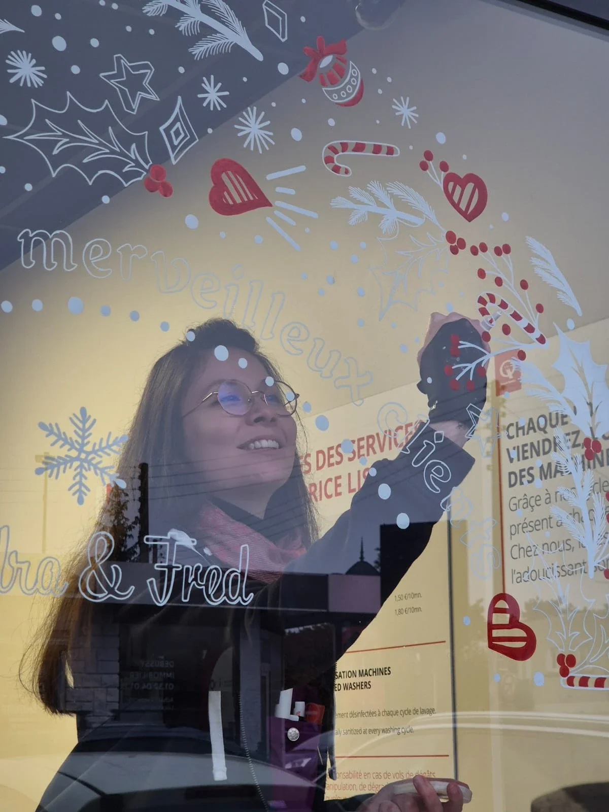 Sandrine Daniel réalise à la main une peinture sur vitrine de Noël représentant une demi-couronne décorative pour une laverie