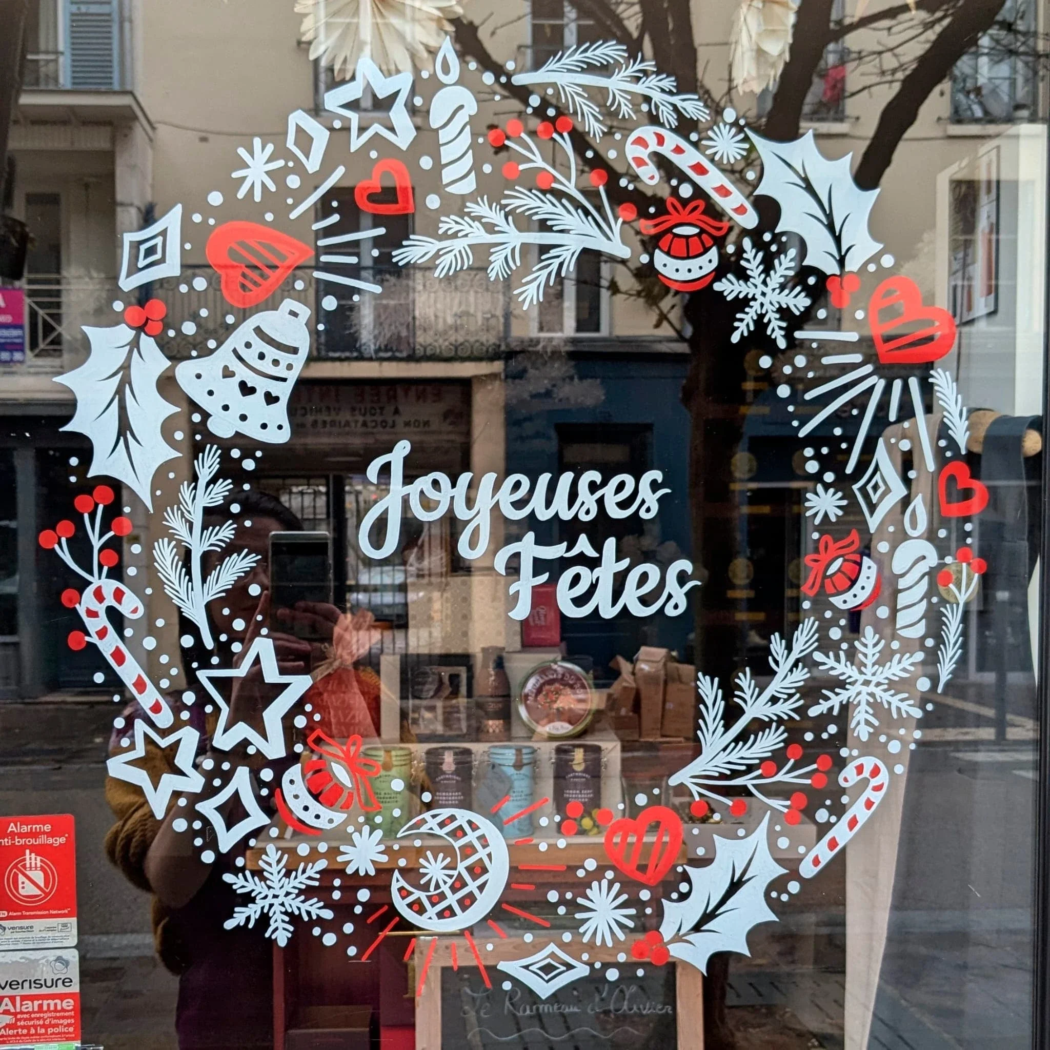 Peinture sur vitrine représentant une couronne de Noël peinte à la main avec houx, cloches, étoiles, baies et branches de pin pour l'épicerie fine Le Rameau d'Olivier à Saint-Germain-en-Laye (78)