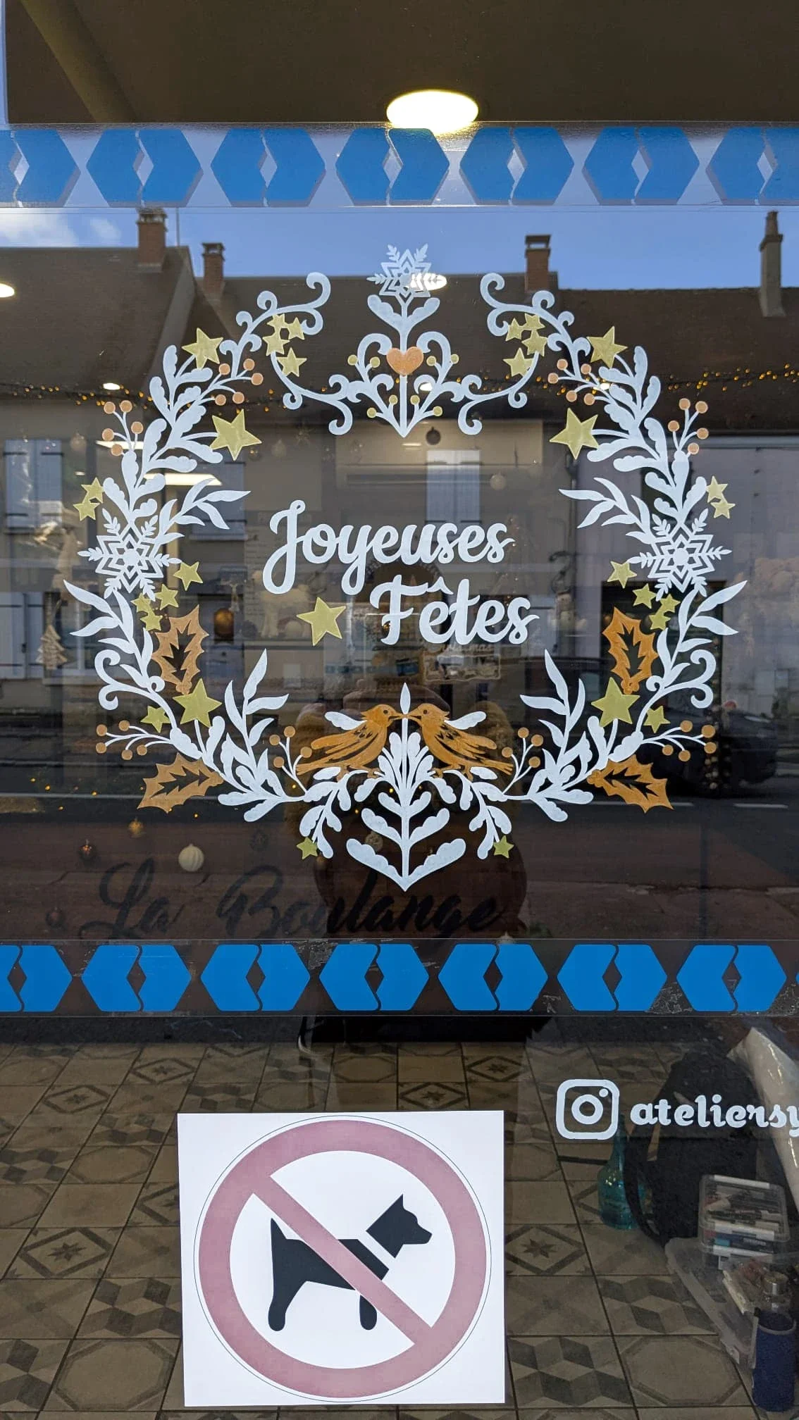 Peinture sur vitrine de Noël pour la boulangerie La Boulange à La Chapelle-en-Serval - une couronne sur la porte avec entrelacs de végétaux et oiseaux. Coloris blanc, doré et cuivré
