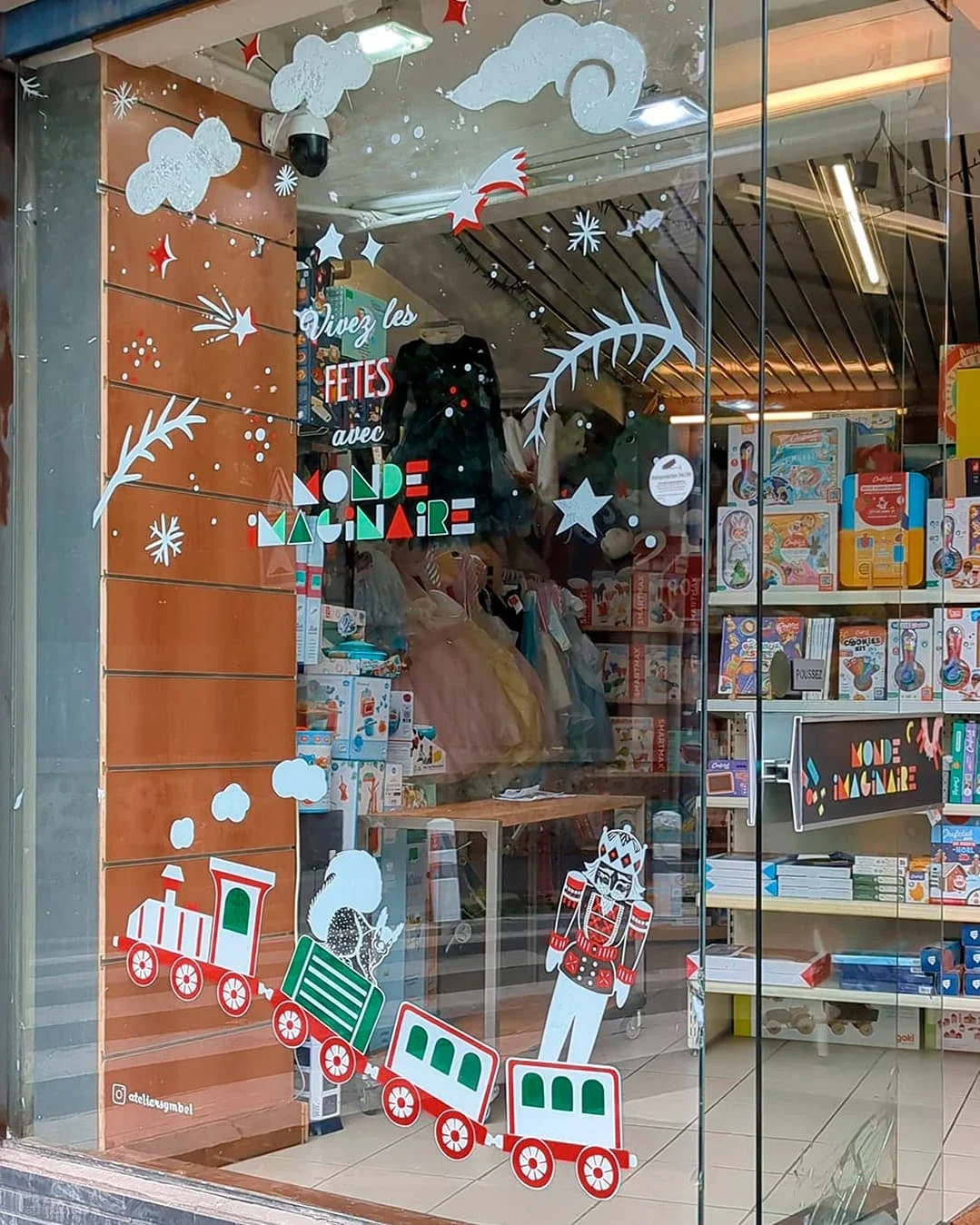 Peinture sur vitrine de Noël sur-mesure pour une boutique de jouets à Houilles (78), avec lettrage et illustrations : branches, étoiles, train d'autrefois, casse-noisette et écureuils
